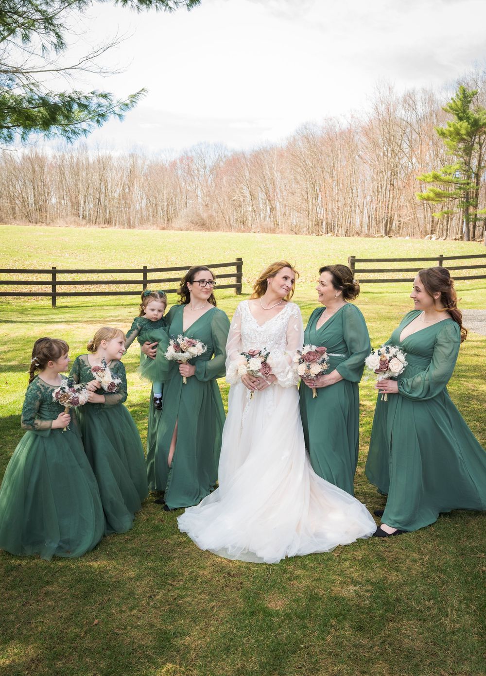 Bride with bridesmaids in emerald green gowns holding bouquets on a green lawn at outdoor wedding.