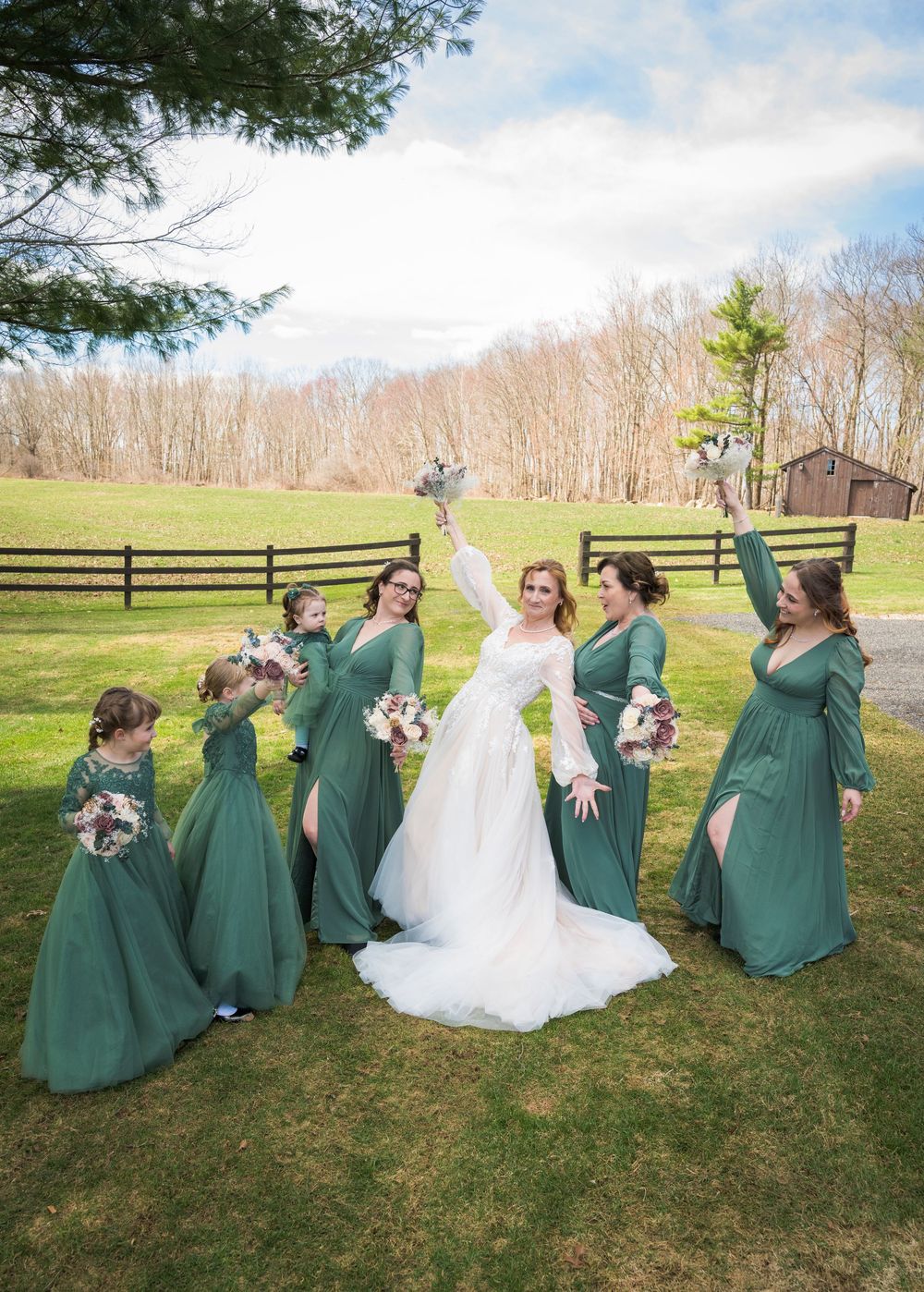 Bride in white gown celebrates with bridesmaids in emerald green dresses on green lawn at outdoor wedding venue.