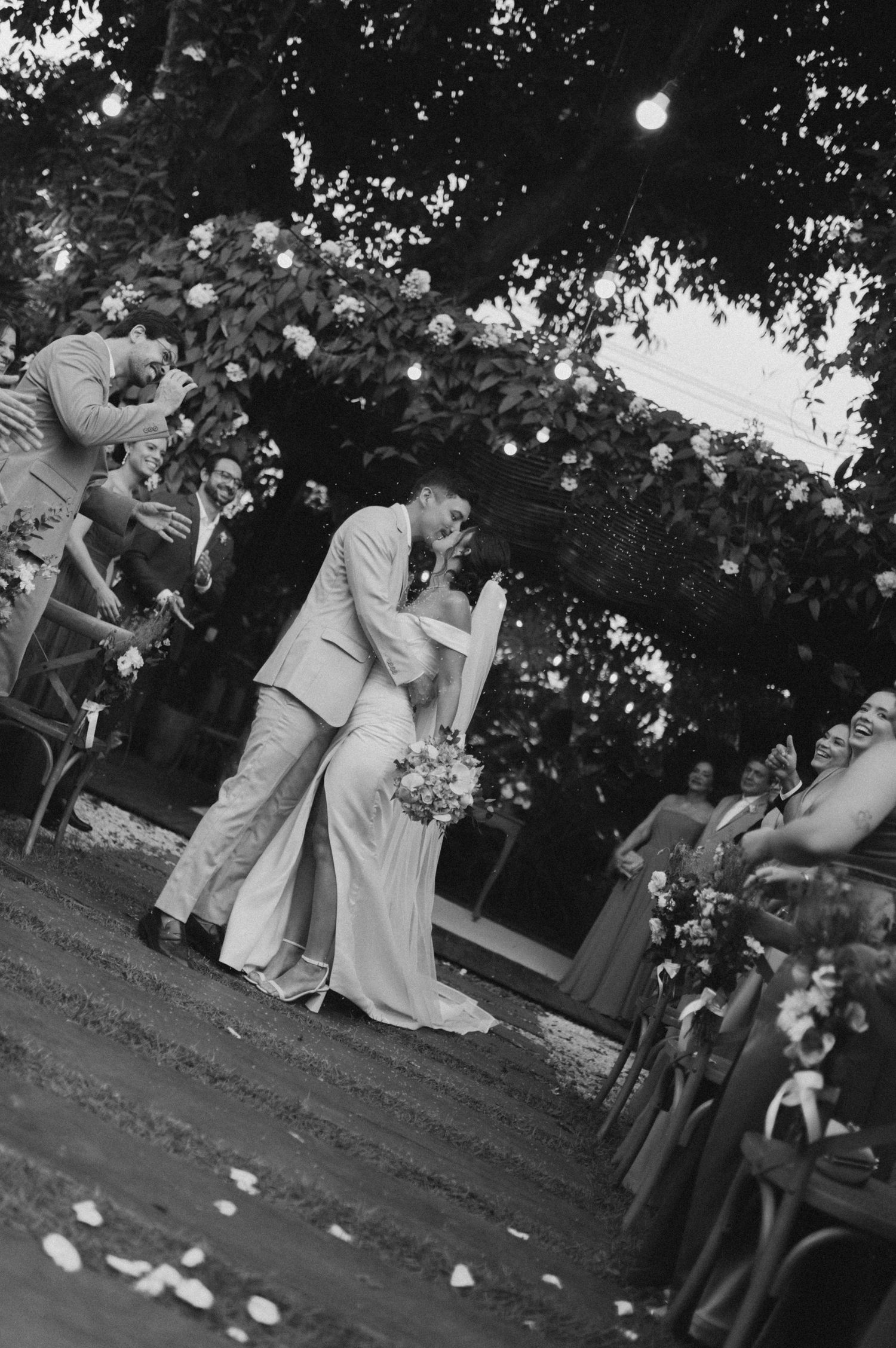 Black and white wedding scene with flower petals falling during an outdoor garden ceremony.
