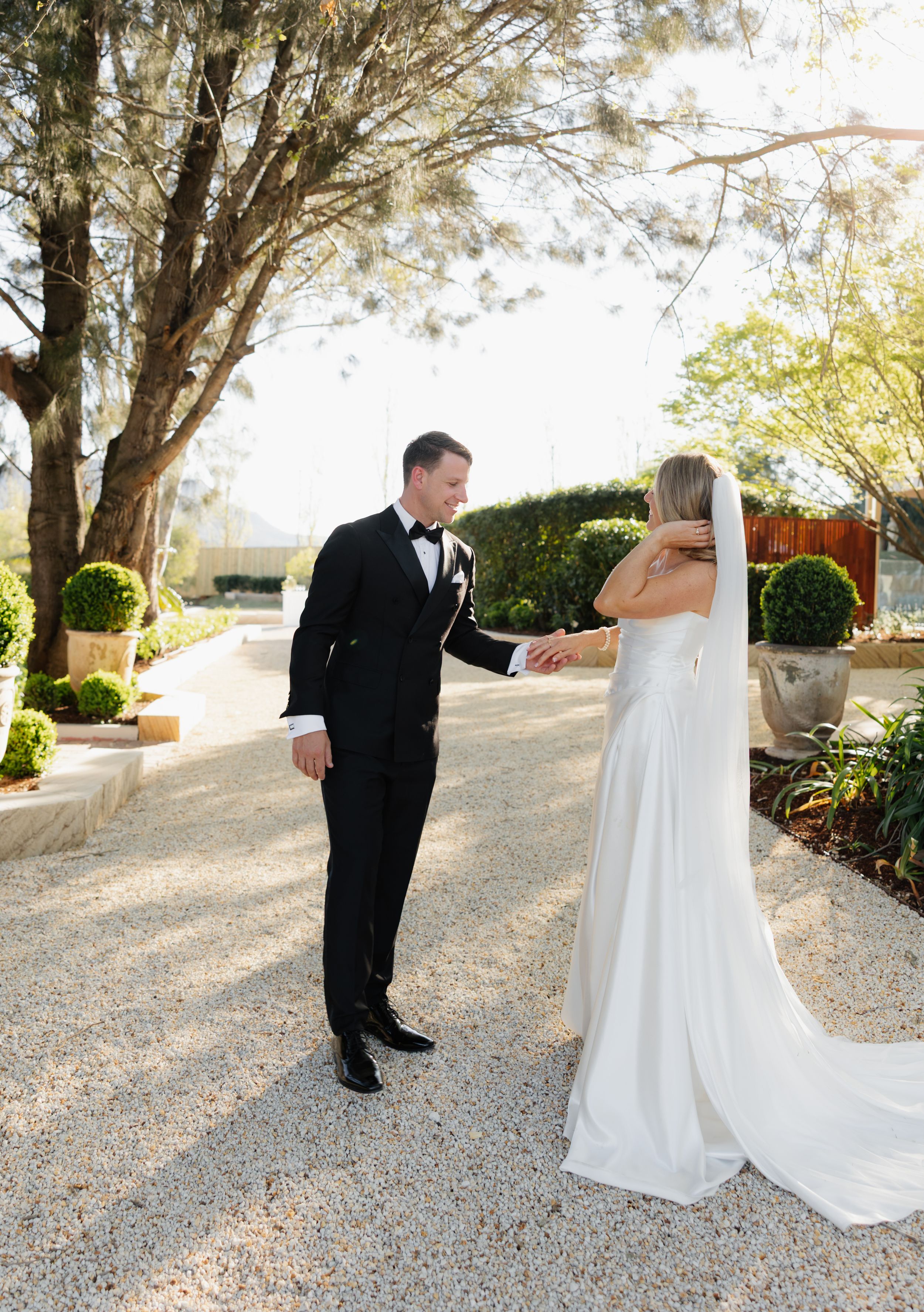 A bride and groom share their first look on a gravel path surrounded by lush garden landscaping on their wedding day.
