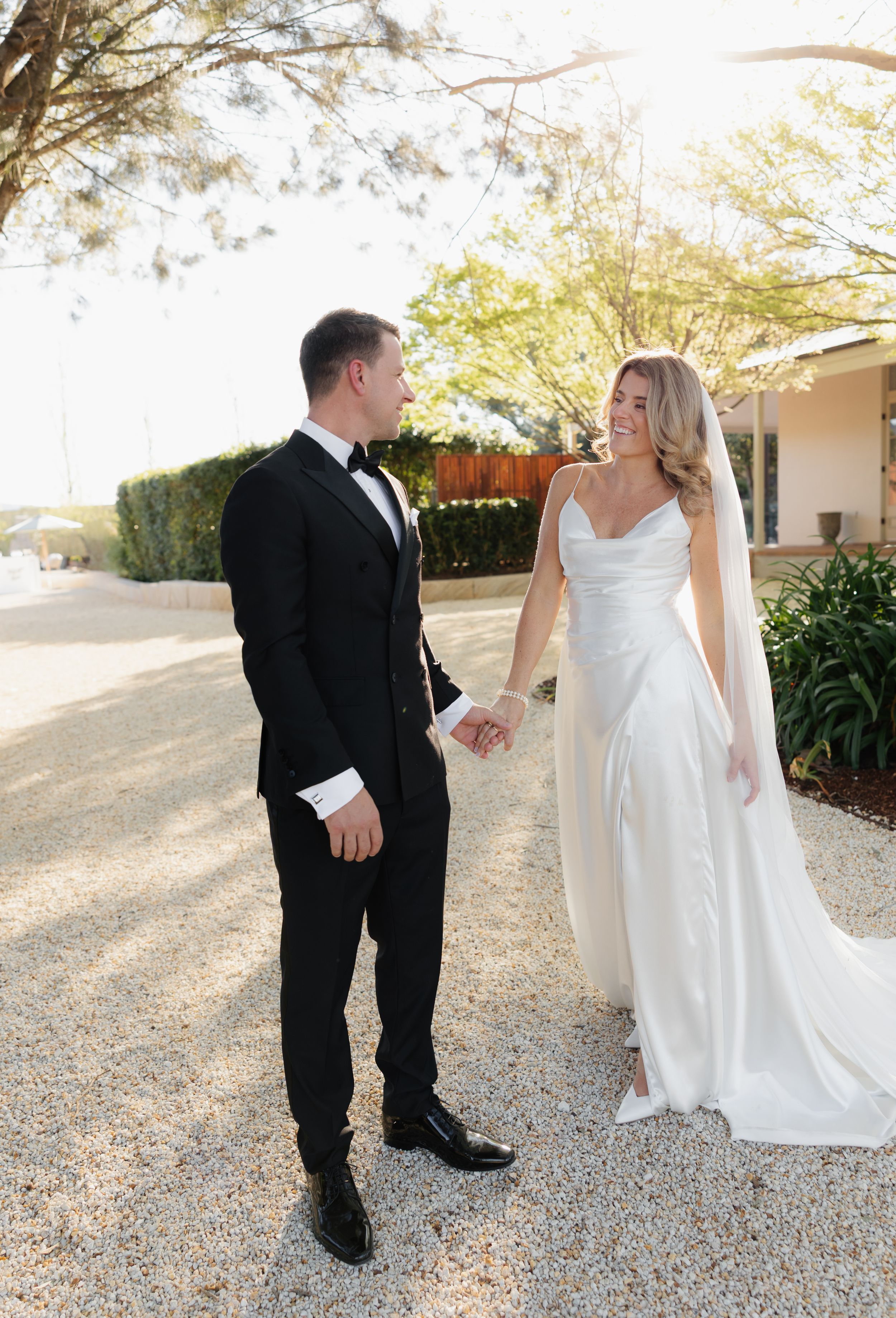 A couple in wedding attire gazes at each other while holding hands on a sunlit garden path.