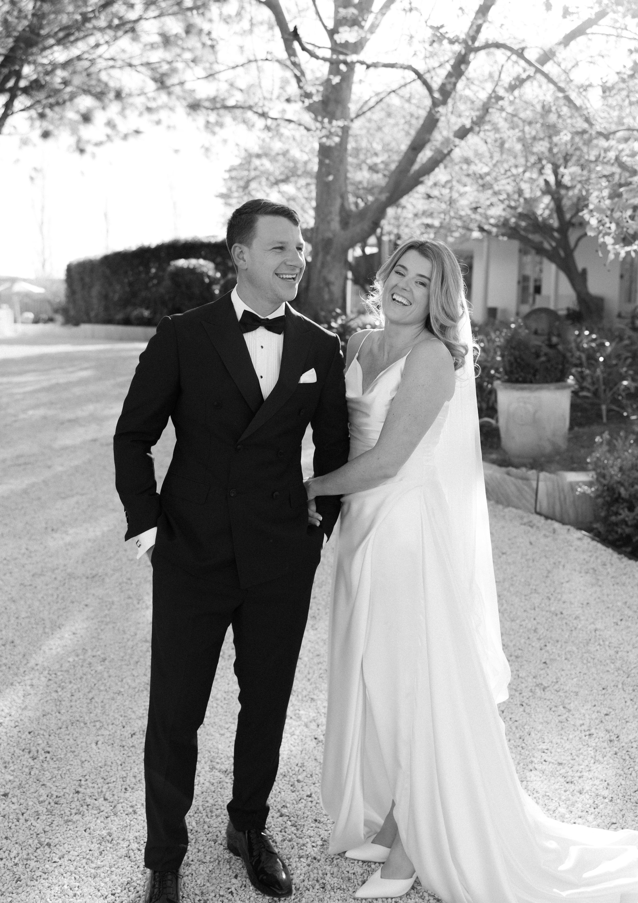 A couple in wedding attire shares a joyful moment while walking together on a tree-lined path.