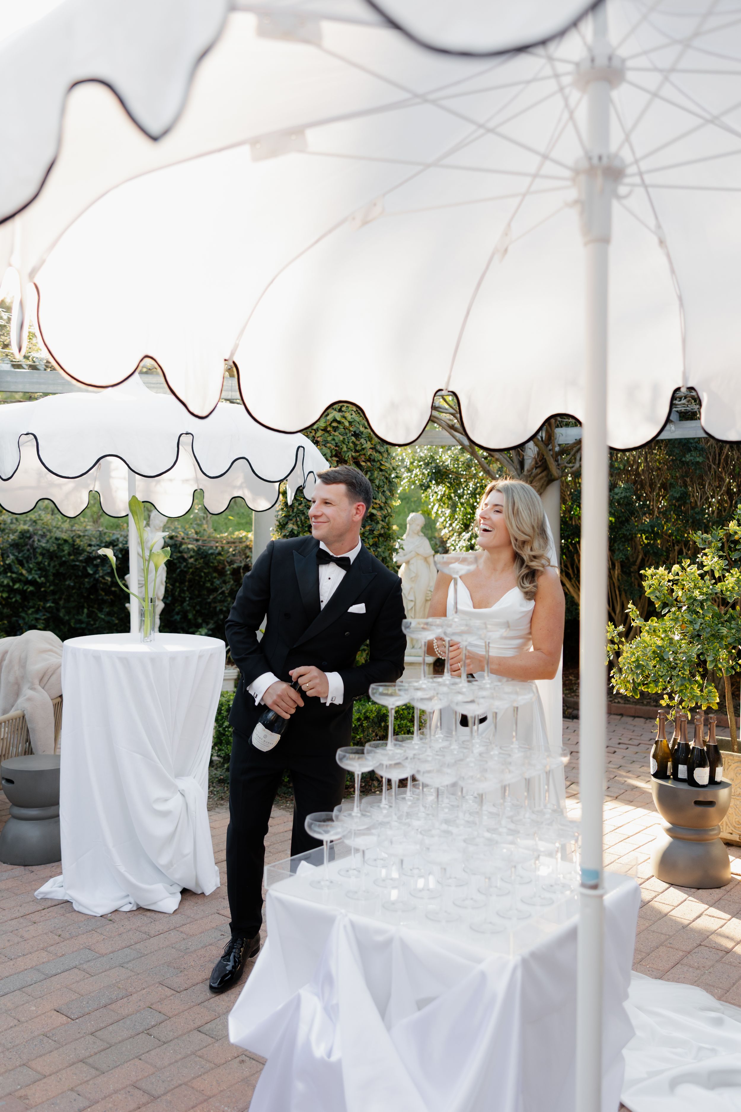 A couple shares a laugh at their elegant outdoor wedding reception under a white scalloped canopy with champagne display.