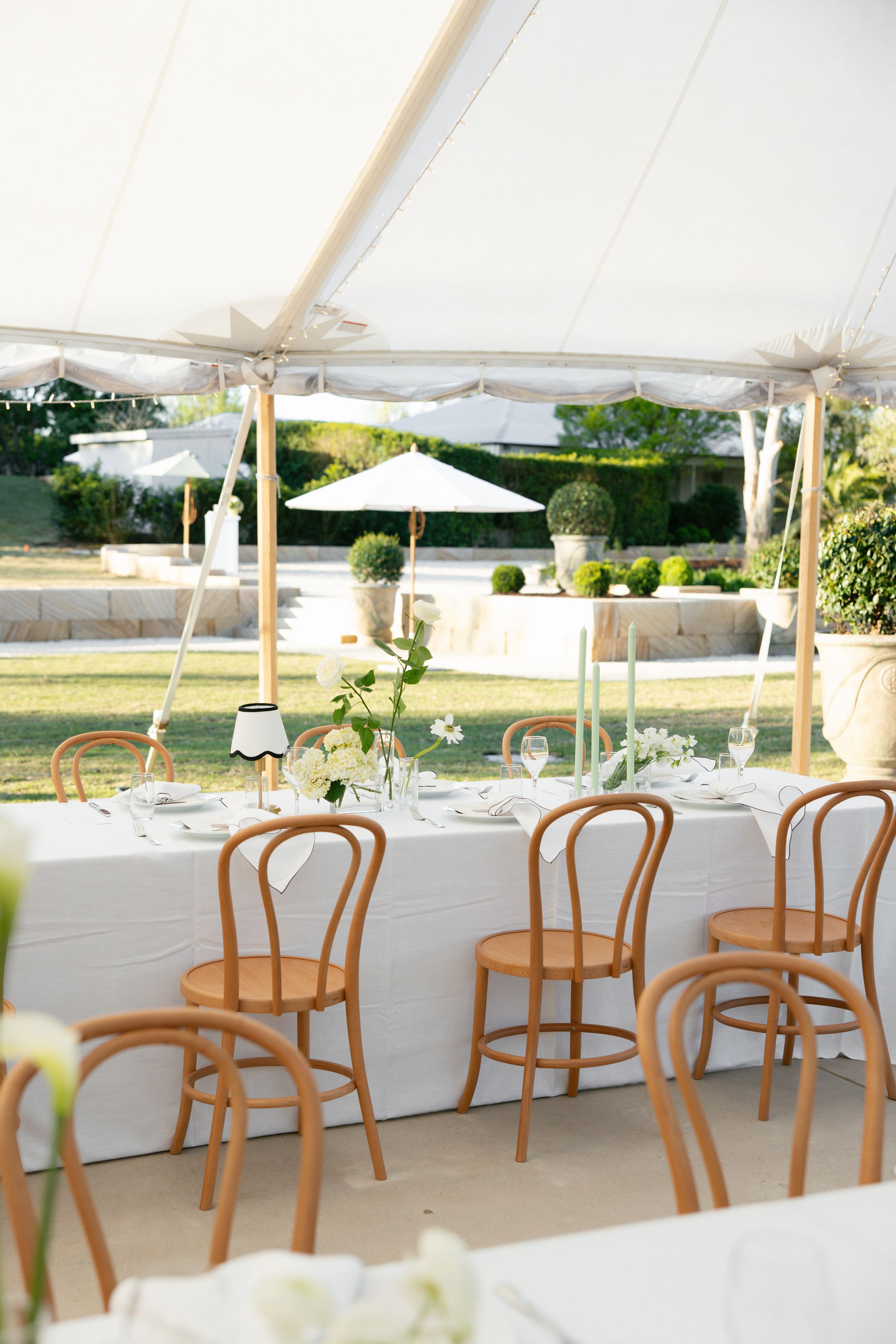 Elegant wedding reception setup with long white tables and wooden bentwood chairs under a white tent canopy.