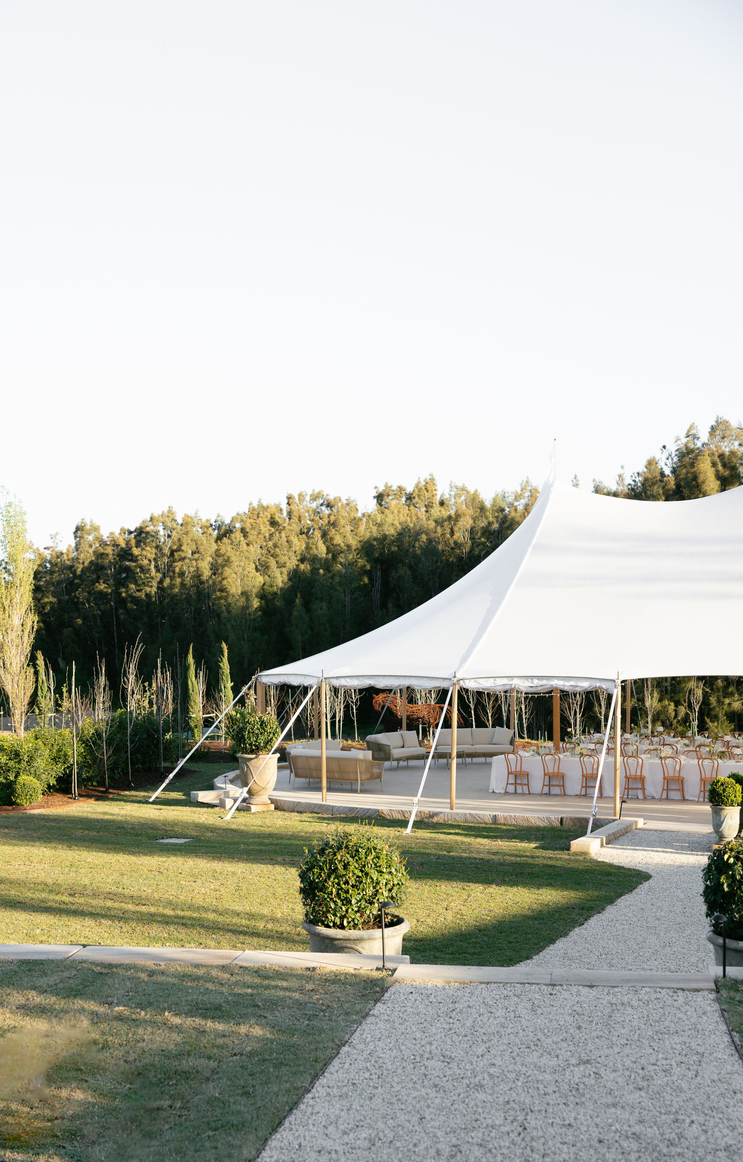 White wedding tent setup on a gravel area surrounded by green lawn and trees during golden hour.