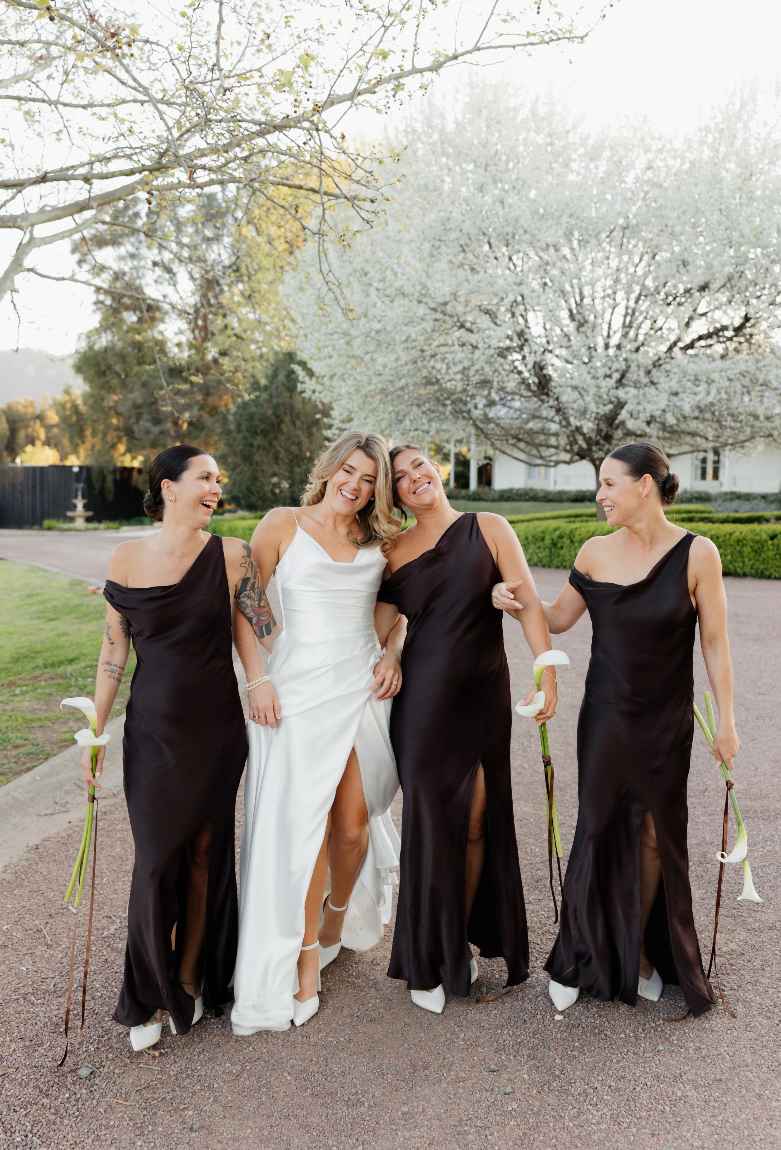 Wedding party in black dresses and white gown pose playfully on gravel path under spring blossoms.