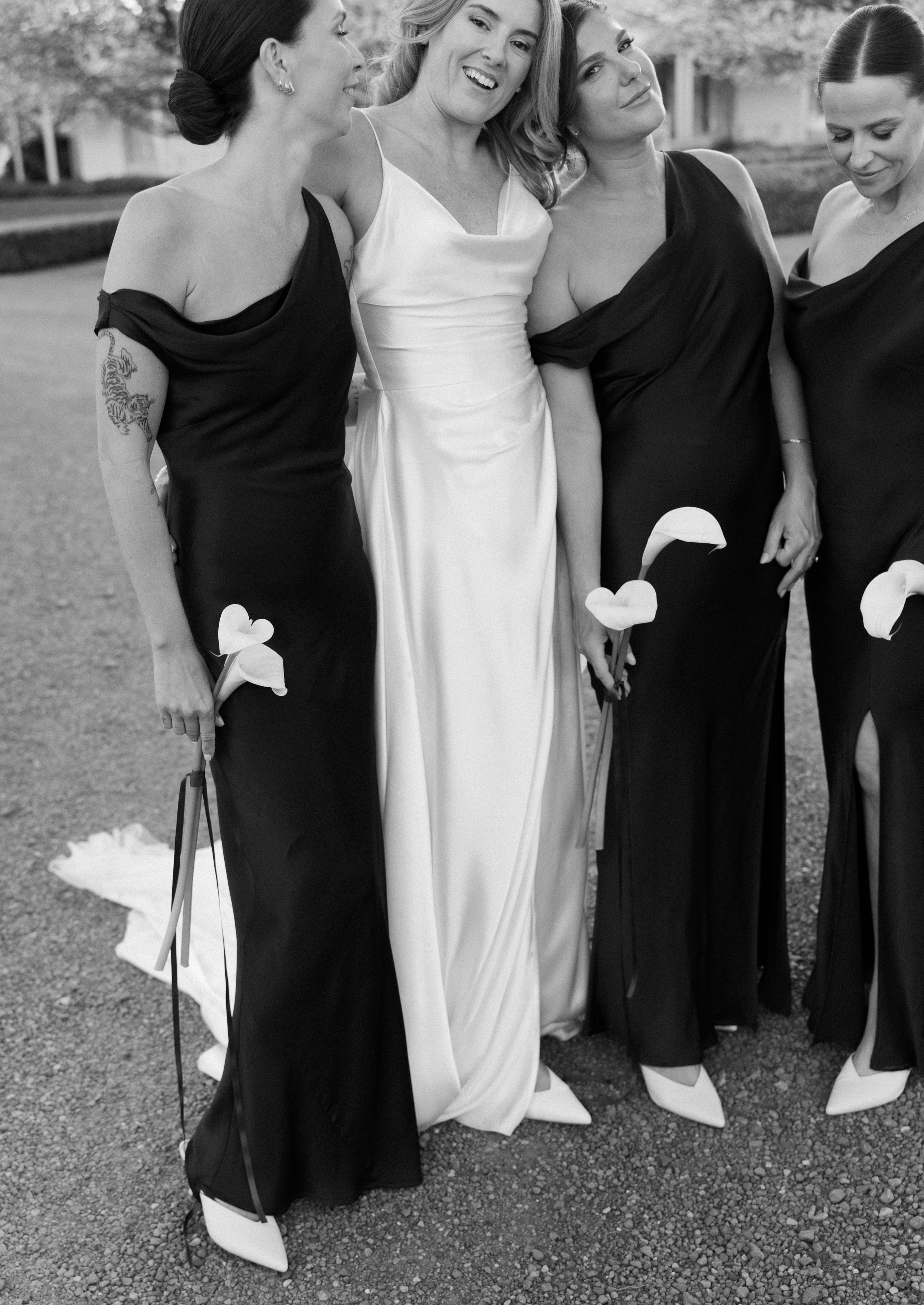 A wedding party in black and white dresses poses together outdoors, showing their shoes in a playful group photo.
