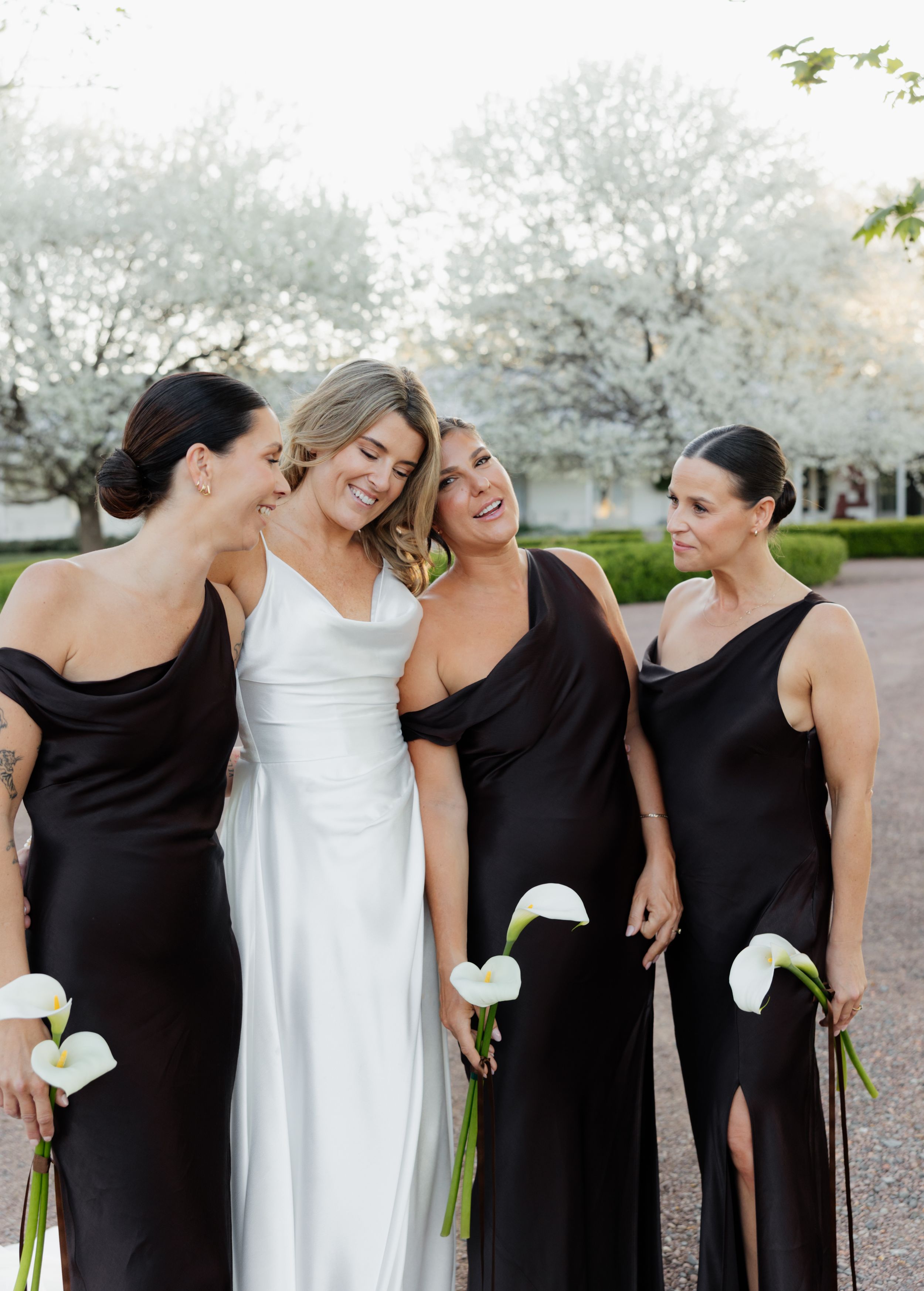 A bridal party wearing black dresses and one white gown pose together in a candid moment against a garden backdrop.