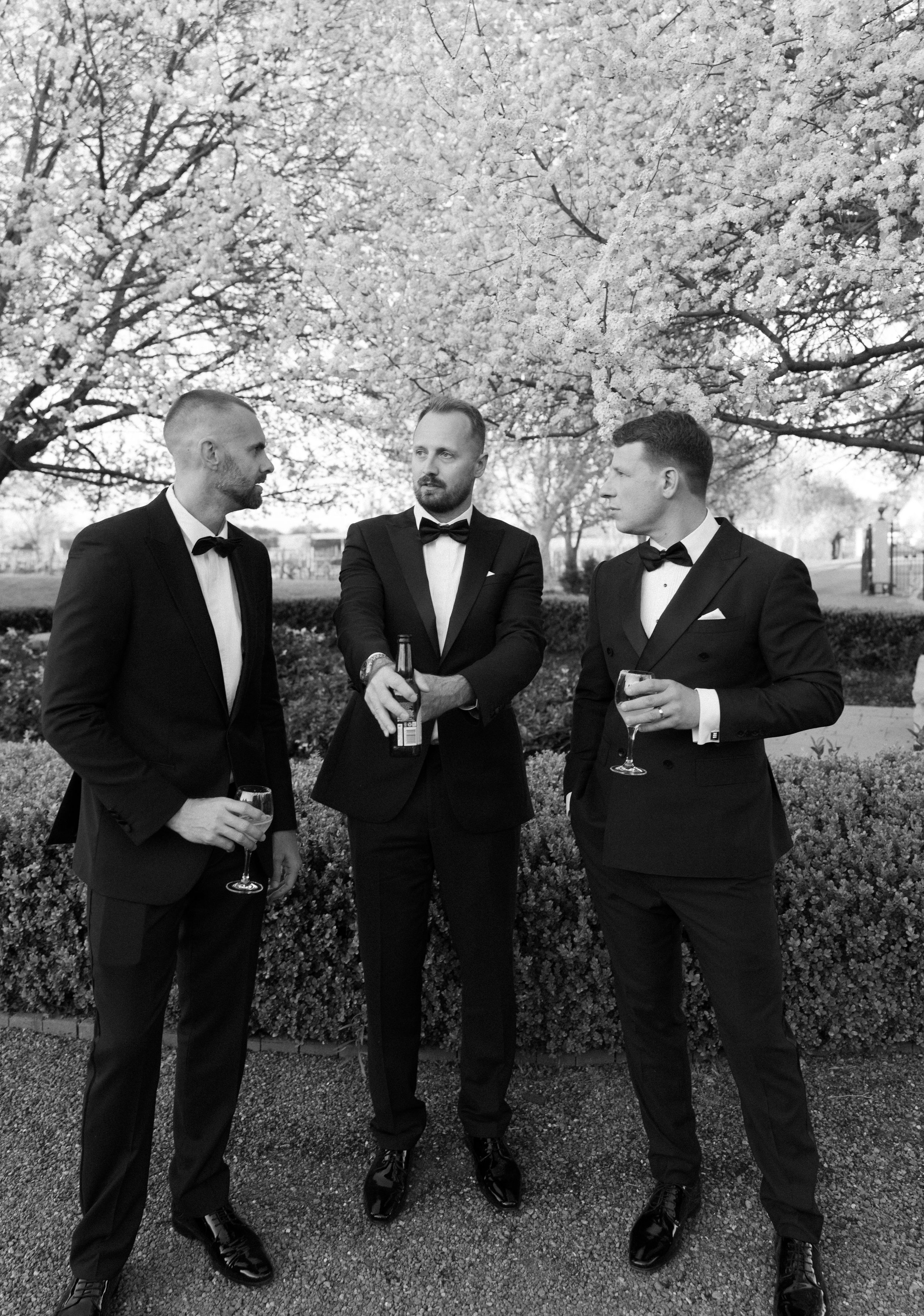 Groomsmen in black tuxedos share drinks and laughs in a series of candid moments under blooming trees.