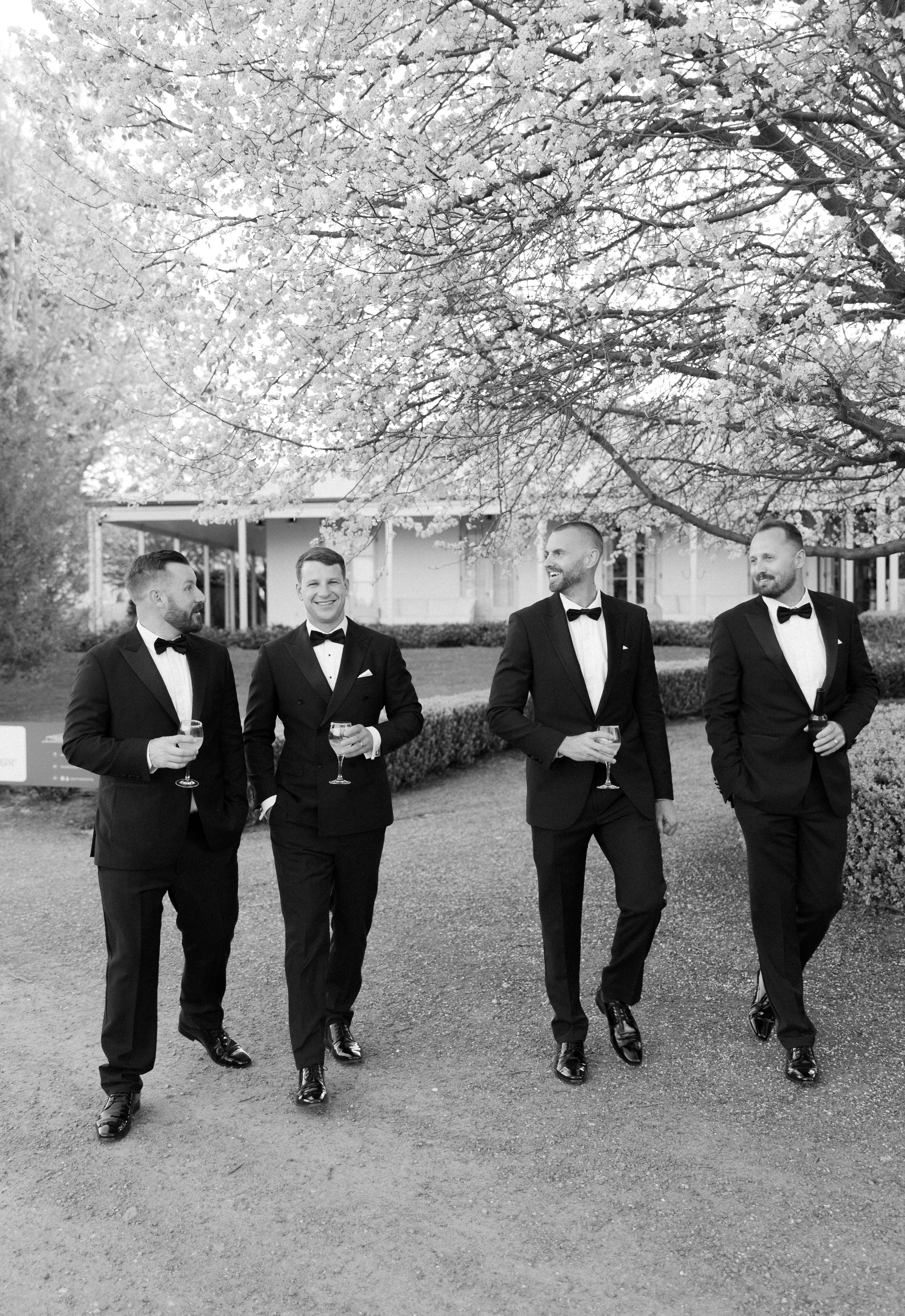 Groomsmen in black tuxedos jump and pose playfully under blooming cherry trees.