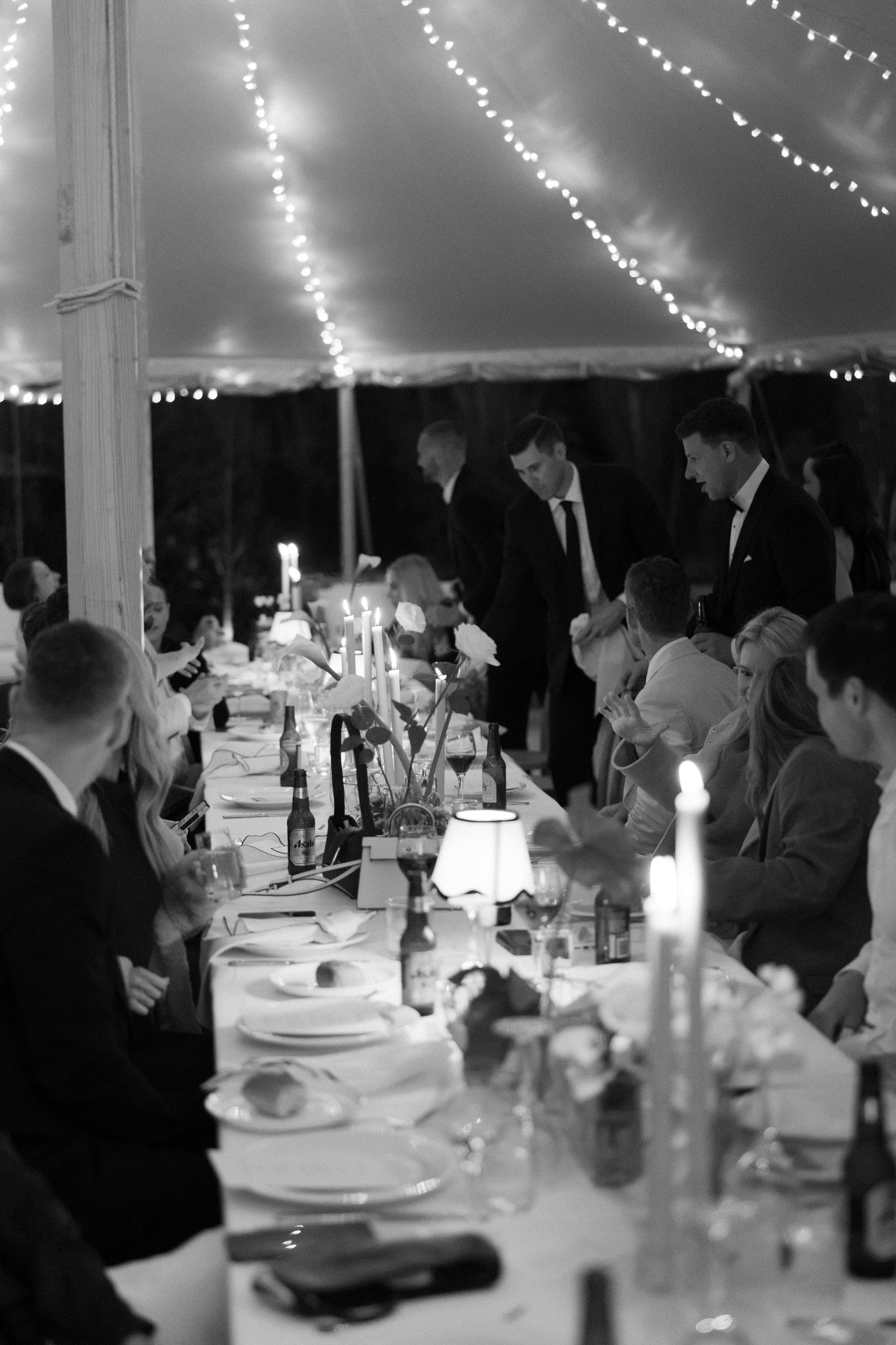 Wedding guests seated at long tables under string lights at an elegant evening reception dinner.