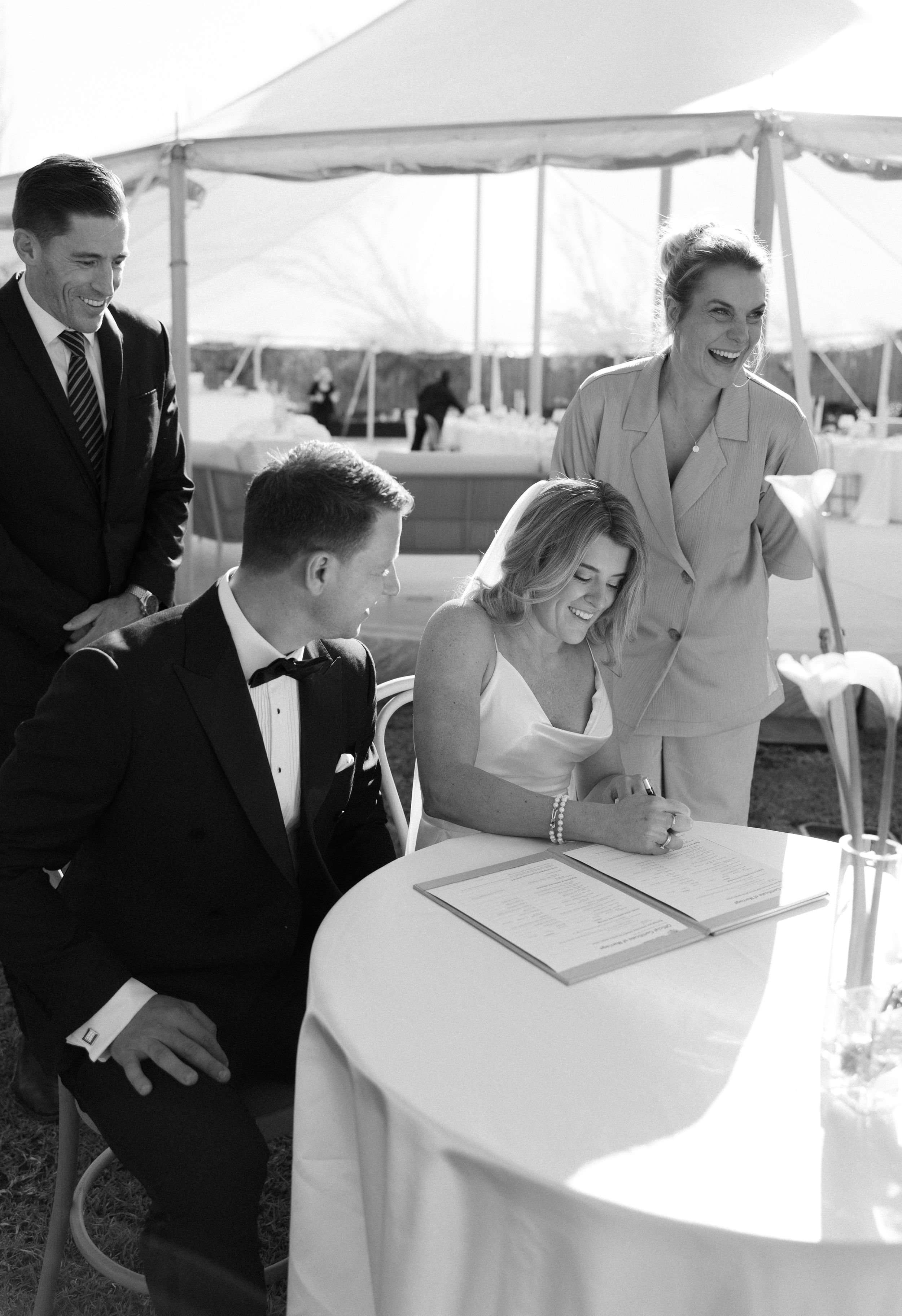Black and white photo of wedding guests signing the guest book under a white tent at an outdoor reception.