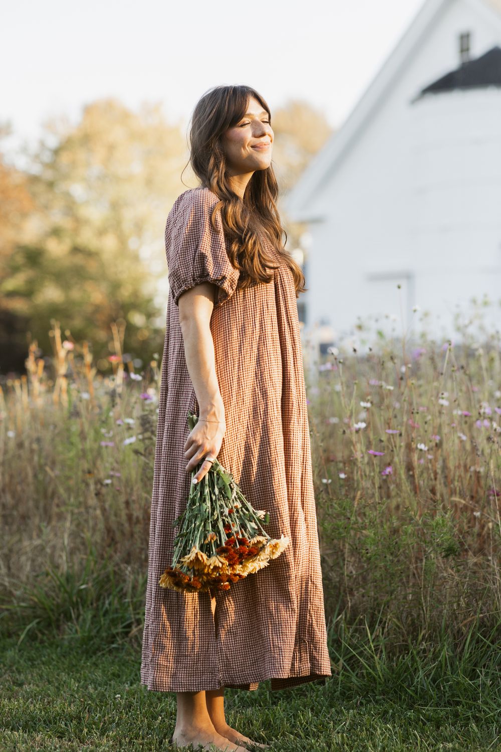 A mother with long wavy brown hair wearing a tan gingham dress holds a bouquet of orange daisies in a field in front of Saltonstall Farm NH.