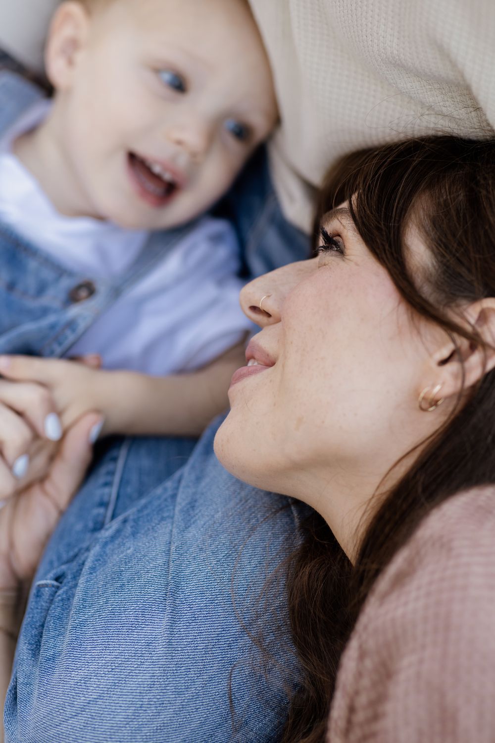 A mother with dark hair wearing gingham gazes adoringly at her blonde toddler son wearing denim overalls while laying on her husband's lap.