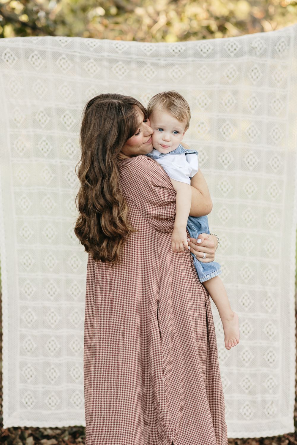 A mother with long brown hair wearing a gingham dress embraces her toddler son in front of a cream crochet blanket for family photos in NH.