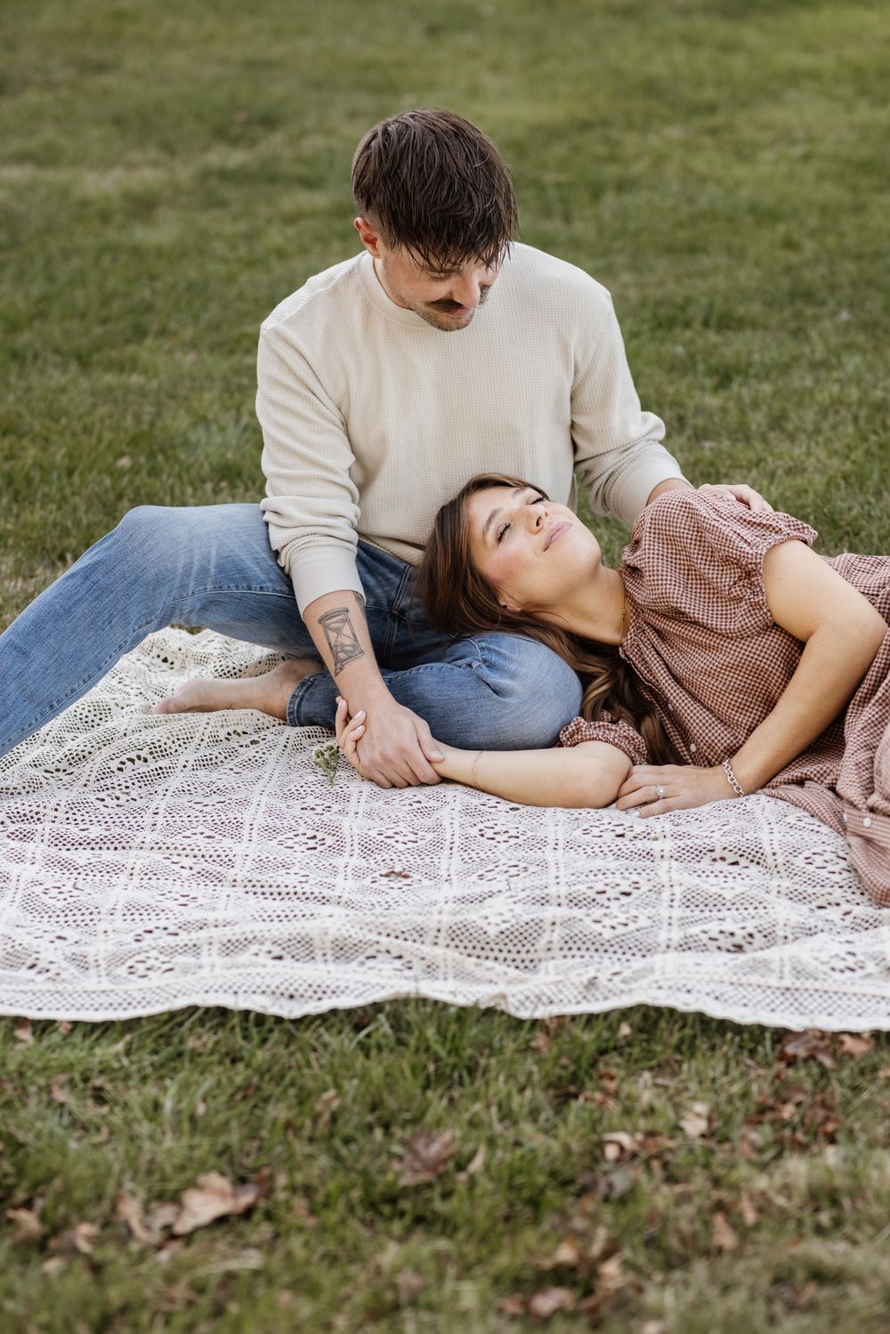A smiling wife wearing a tan gingham dress closes her eyes as she lays on her husband's lap on a cream crochet blanket for family photos.