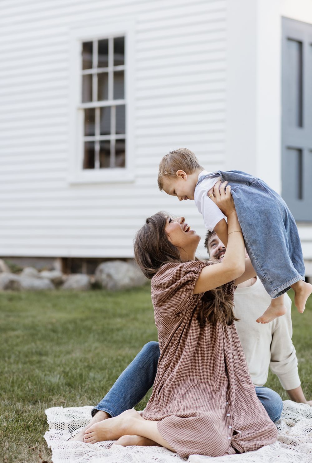 A smiling mother wearing a gingham dress holds her toddler son wearing denim overalls up in the air for family photos in New Hampshire.