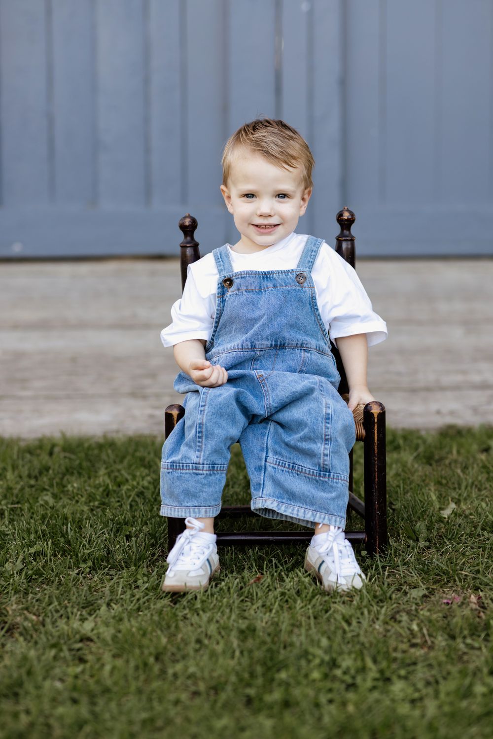 A smiling, blonde hair, blue eyed toddler boy sits on a small wooden chair for family photos at Saltonstall Farm in Stratham, New Hampshire.