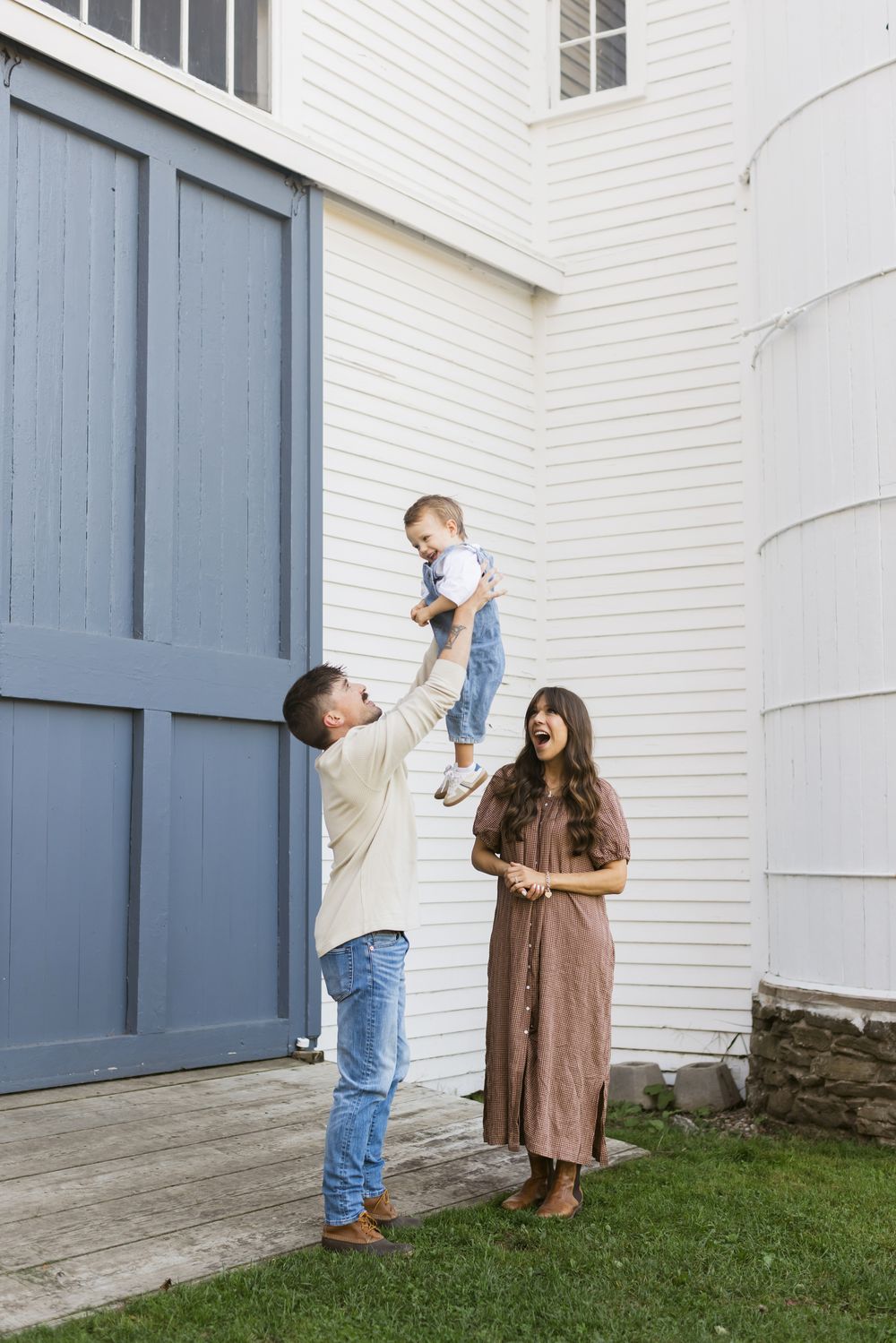 A father wearing blue jeans lifts his son in the air while a smiling mother in a gingham dress looks on in front of a blue barn door in NH.