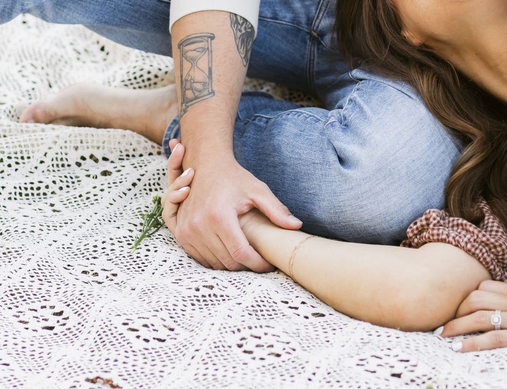 A close up view of a husband with am hourglass tattoo holds his wife's hand on a cream crochet blanket during family photos in New Hampshire