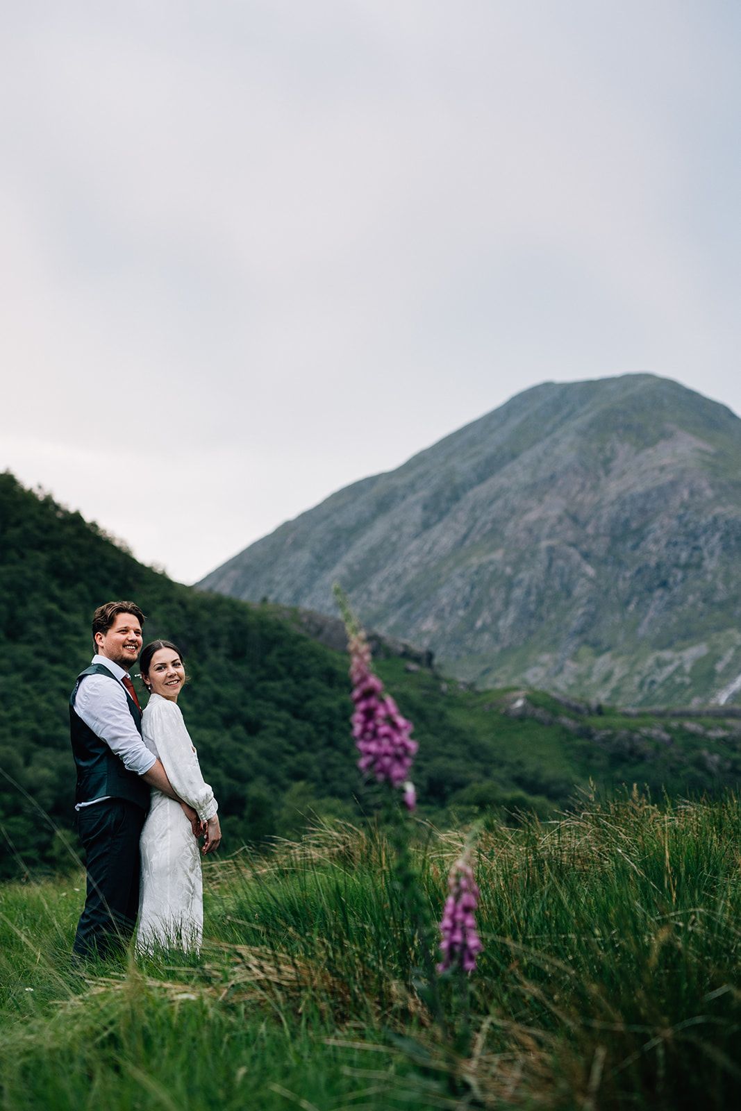 A couple embraces in a romantic moment against the backdrop of majestic mountains and wildflowers in Scotland.