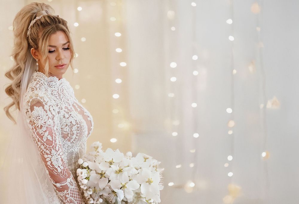 Elegant bride in lace long-sleeve wedding gown holding white floral bouquet against bokeh light backdrop.