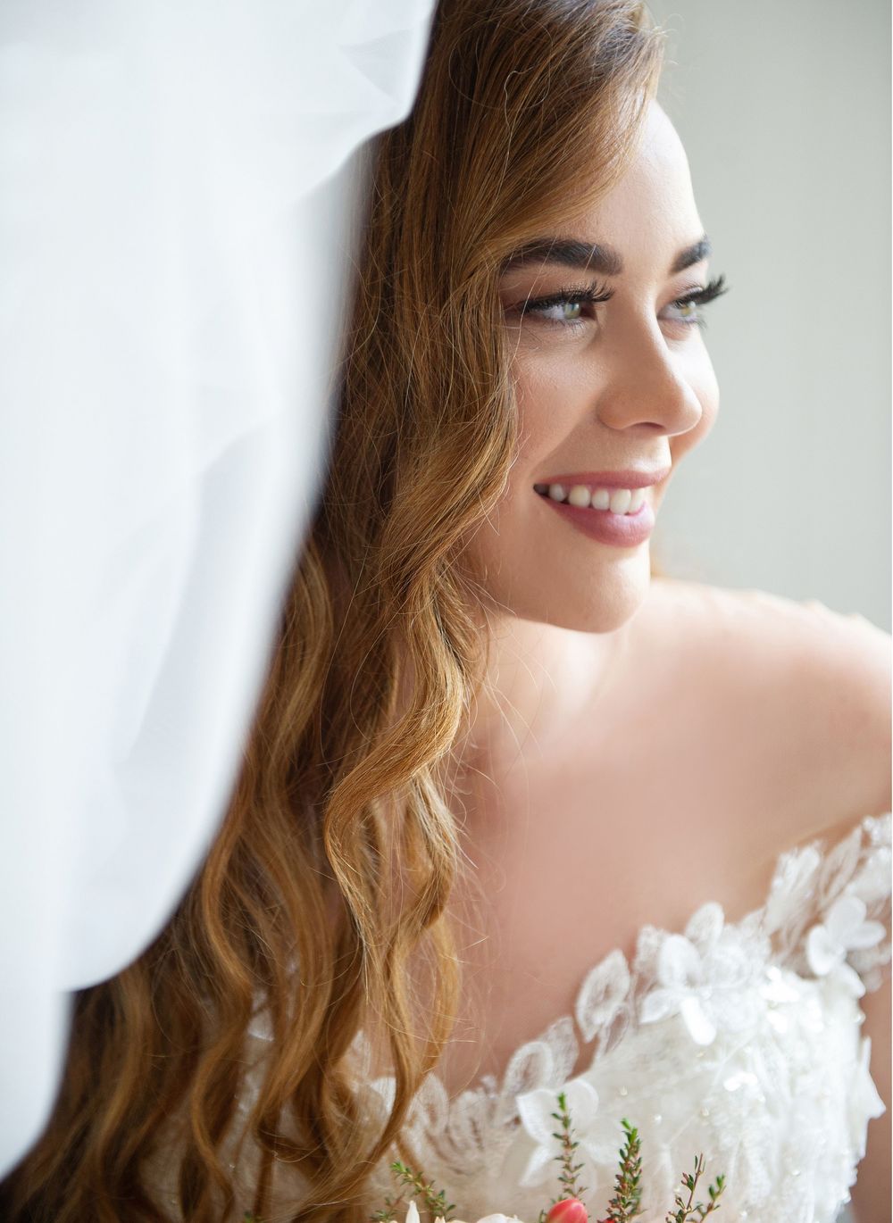Beautiful smiling bride with long wavy hair and floral lace dress holding bouquet beside white veil.