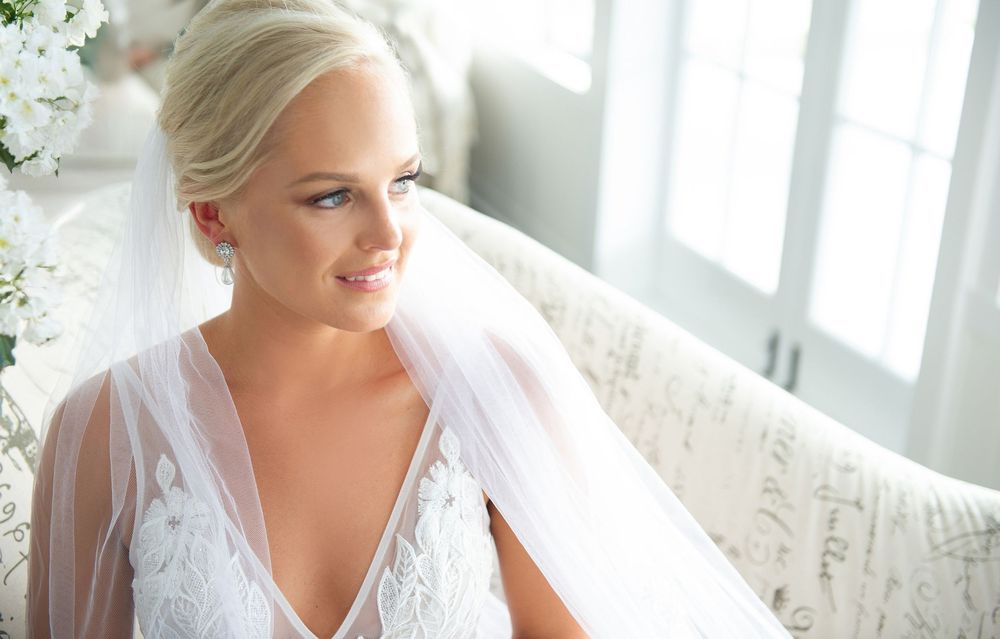 Blonde bride wearing a lace wedding dress and veil sitting on a sofa in soft natural light.