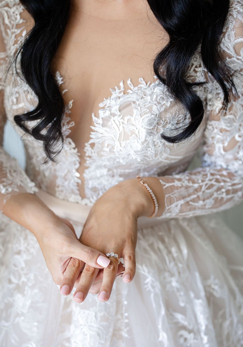 Close-up of bride in deep V-neck lace gown showcasing diamond ring and delicate gold bracelet.