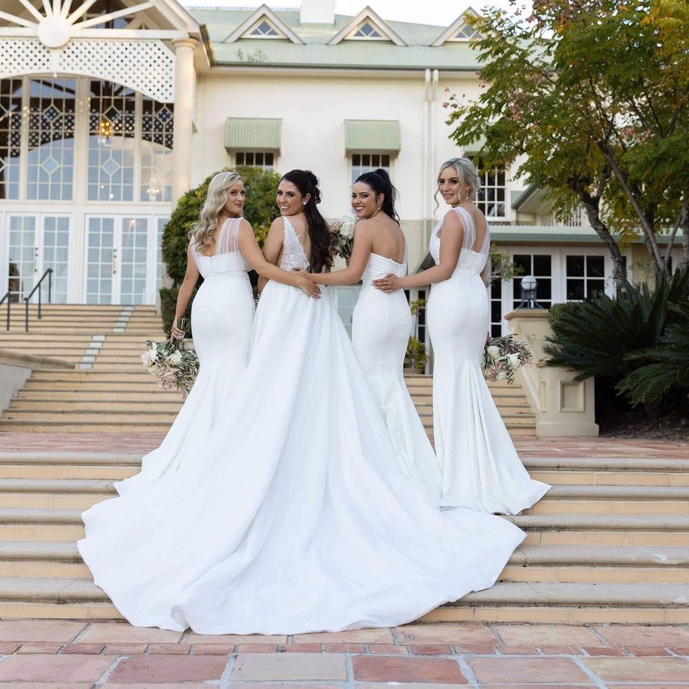 Four bridesmaids in elegant white mermaid gowns pose with their backs to camera on steps of a grand wedding venue.