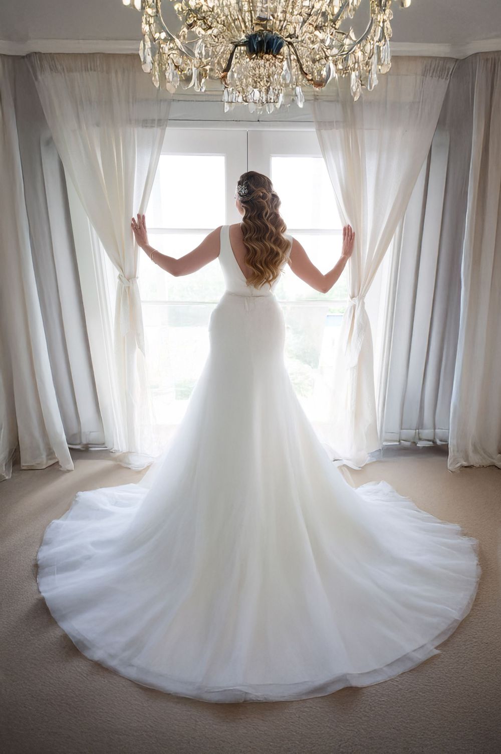 Bride in stunning white ball gown with long train stands before sunlit window beneath crystal chandelier.