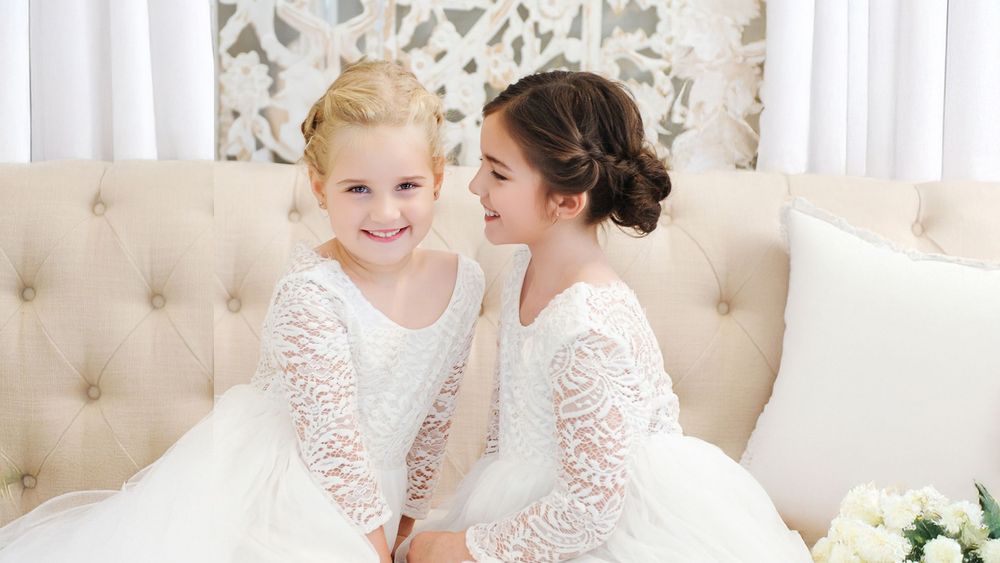 Two young flower girls in white lace dresses sitting together on a tufted cream sofa smiling.