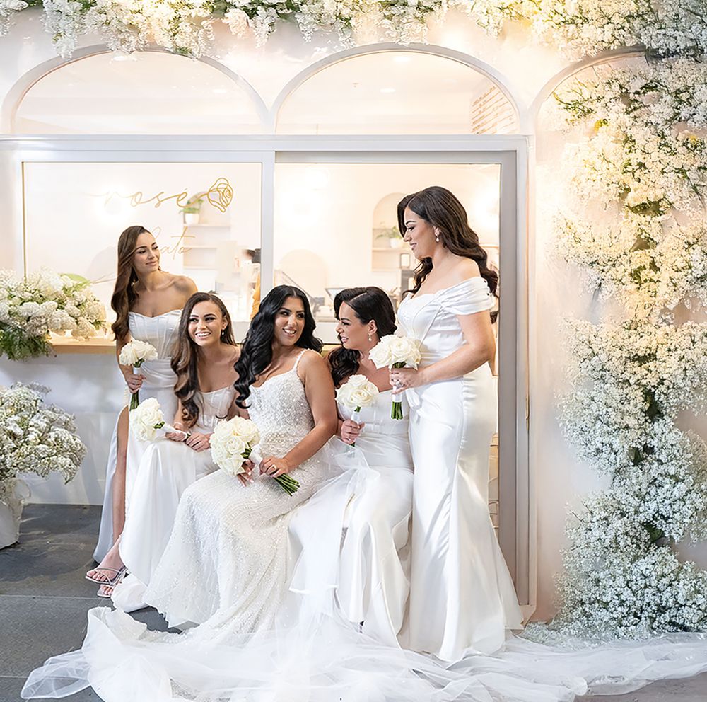 Four bridesmaids in white dresses pose with bride among stunning white floral arch decorations.