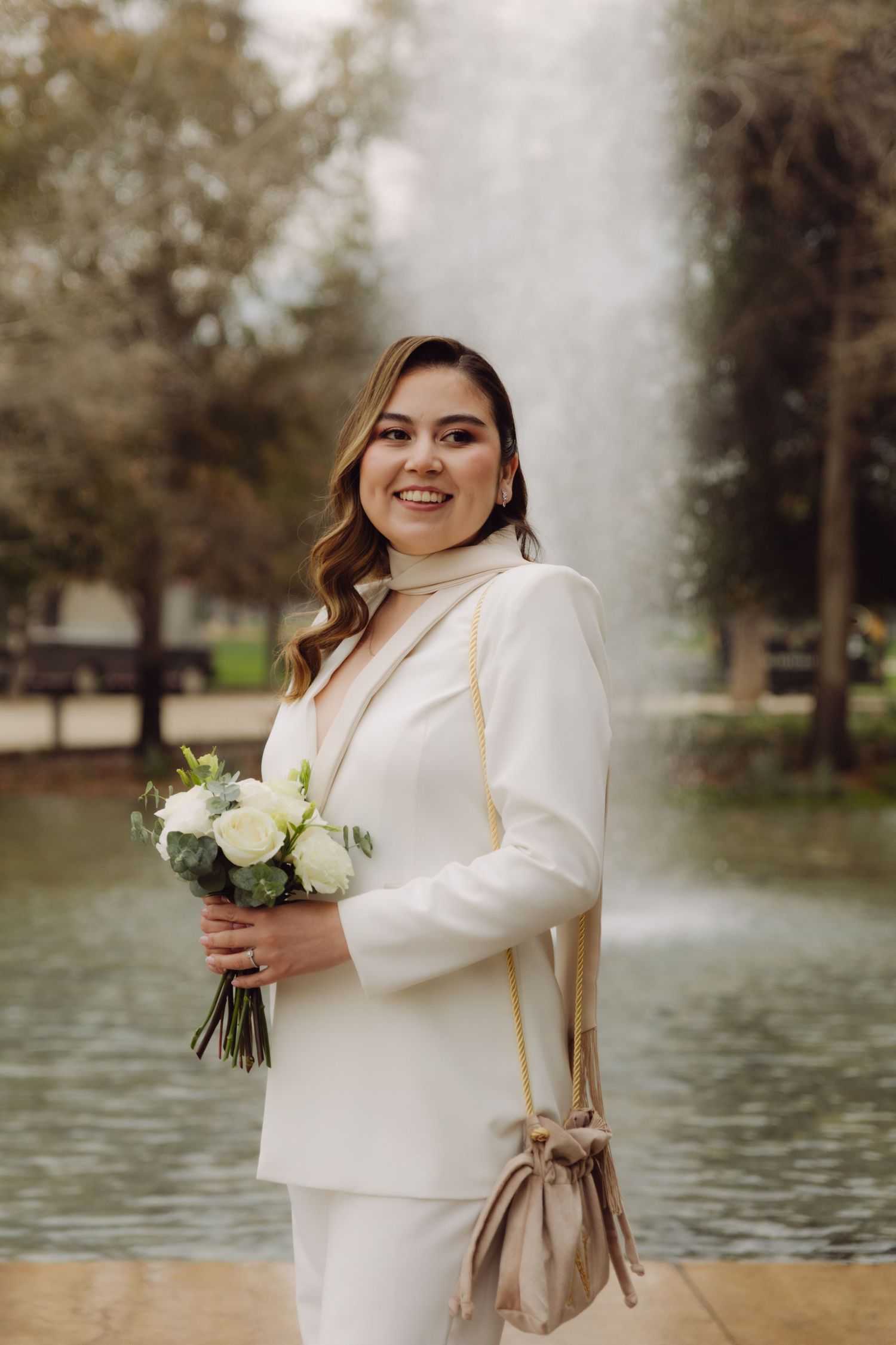 Elegant bride in white turtleneck suit holding white rose bouquet near park fountain.