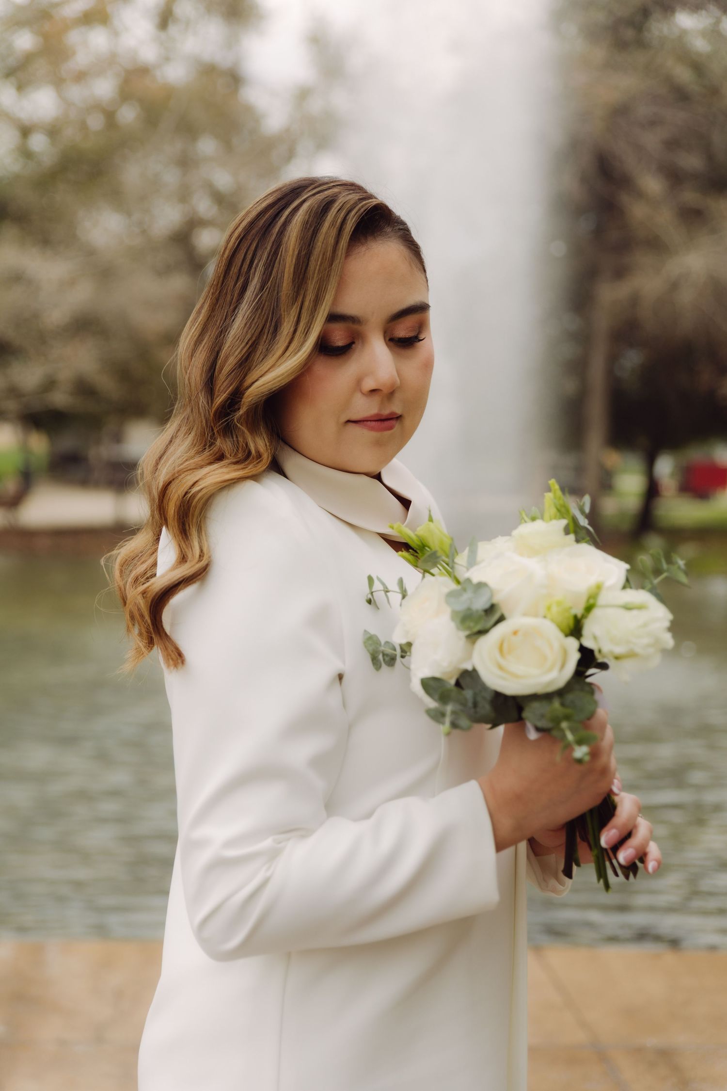 Beautiful bride in white blazer gazing down at white rose bouquet by garden fountain.