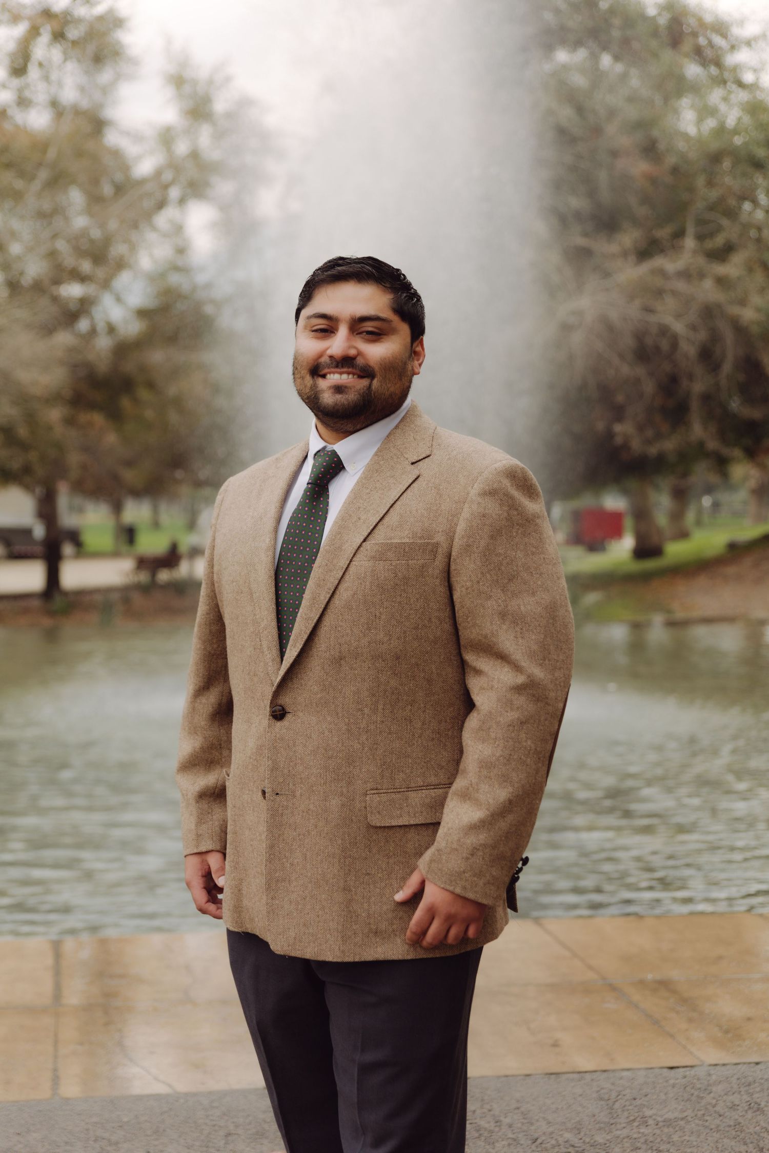Smiling man in tan blazer and green tie posing before a park fountain.