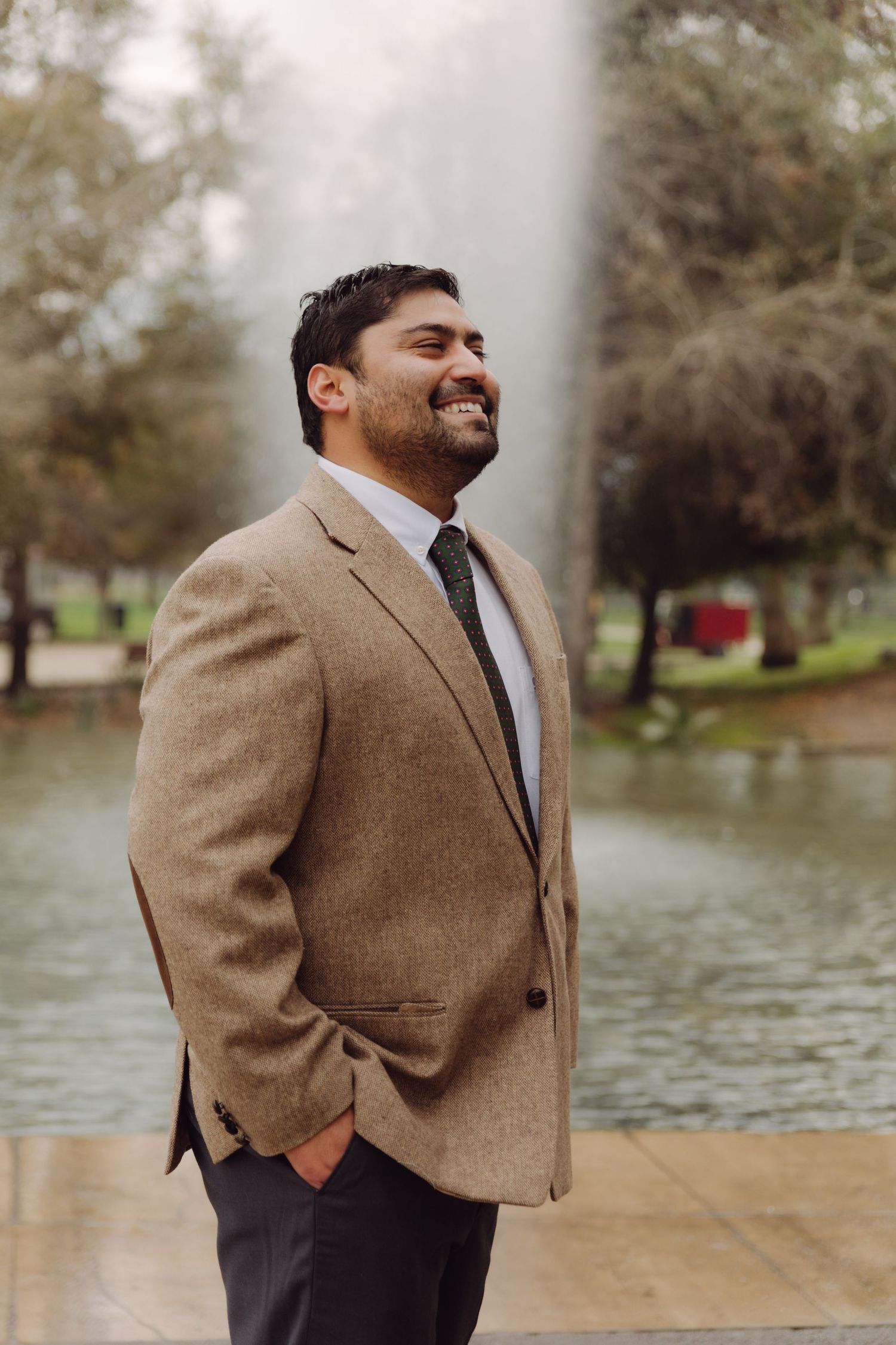 Joyful man in tan suit laughing outdoors near a misty park fountain.
