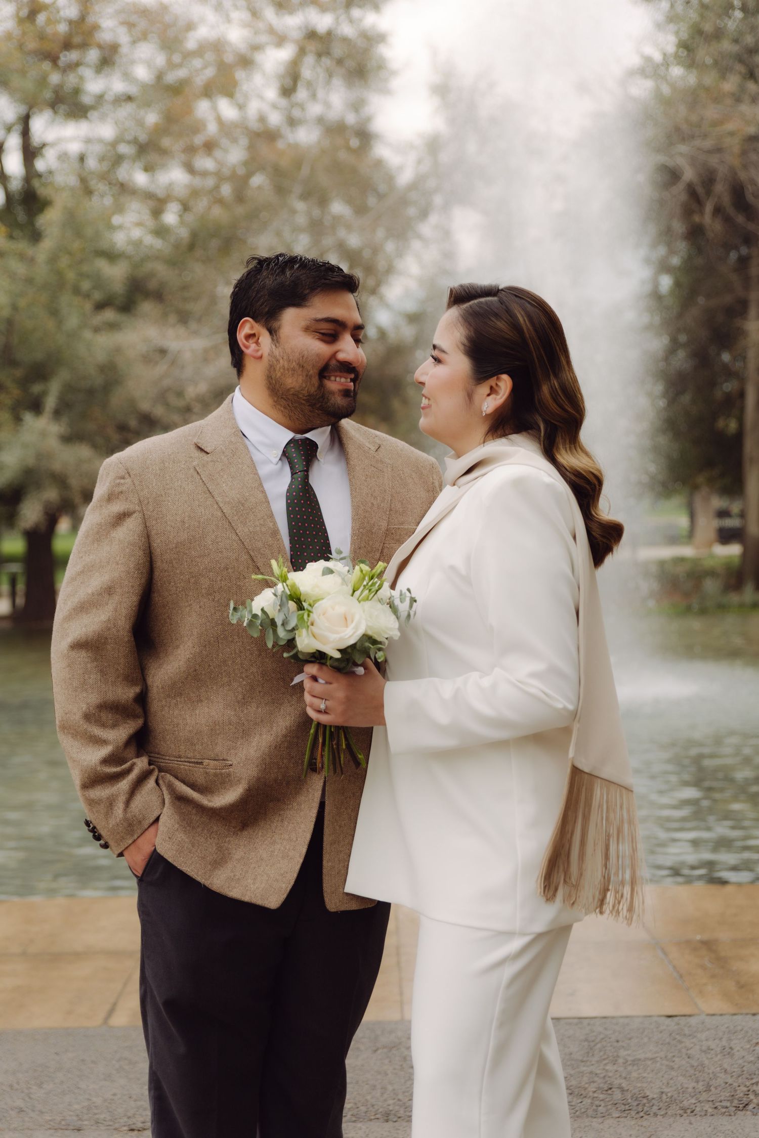 Couple in wedding attire sharing a tender moment by a park fountain.