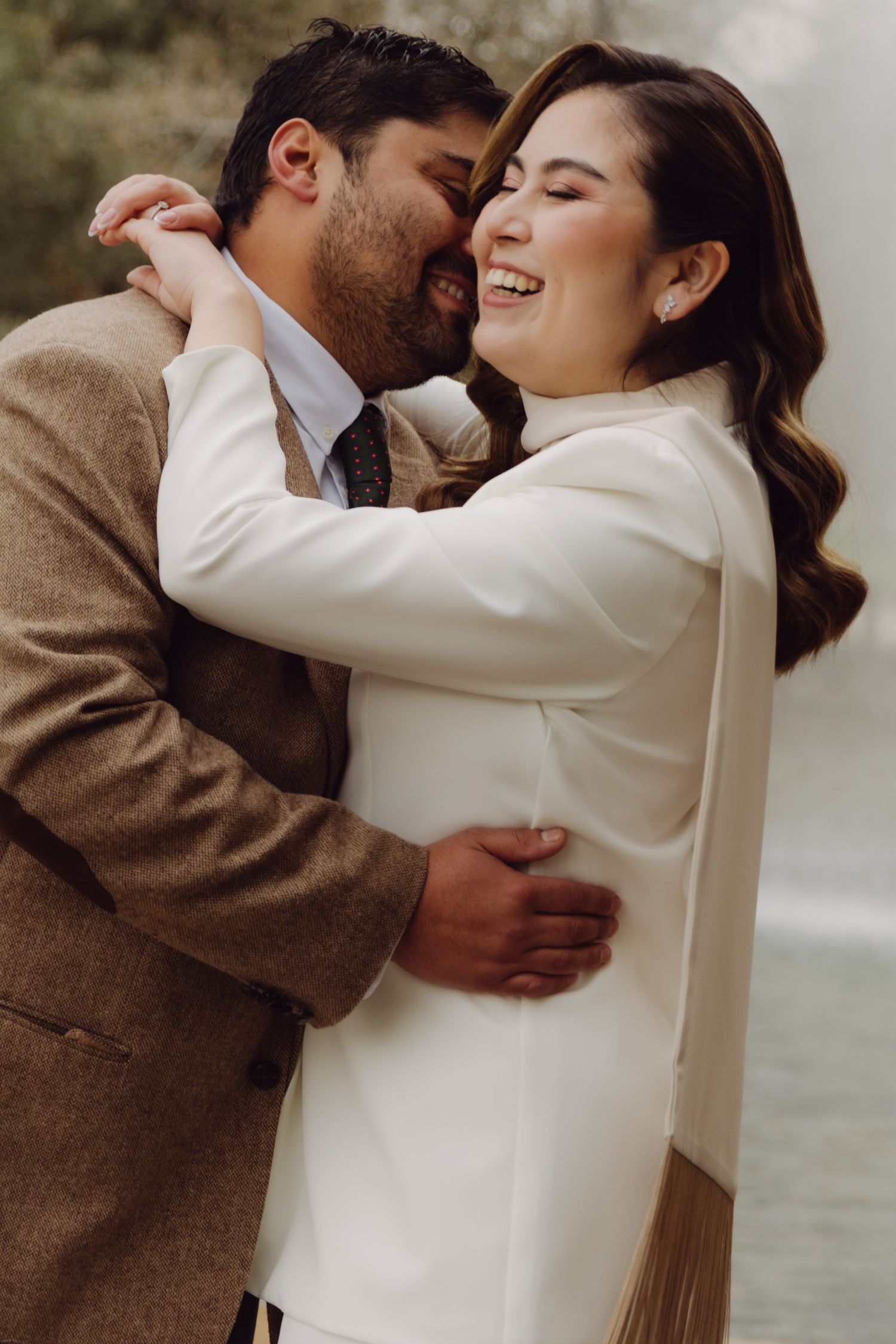 Groom in tan blazer embracing laughing bride in white suit outdoors.