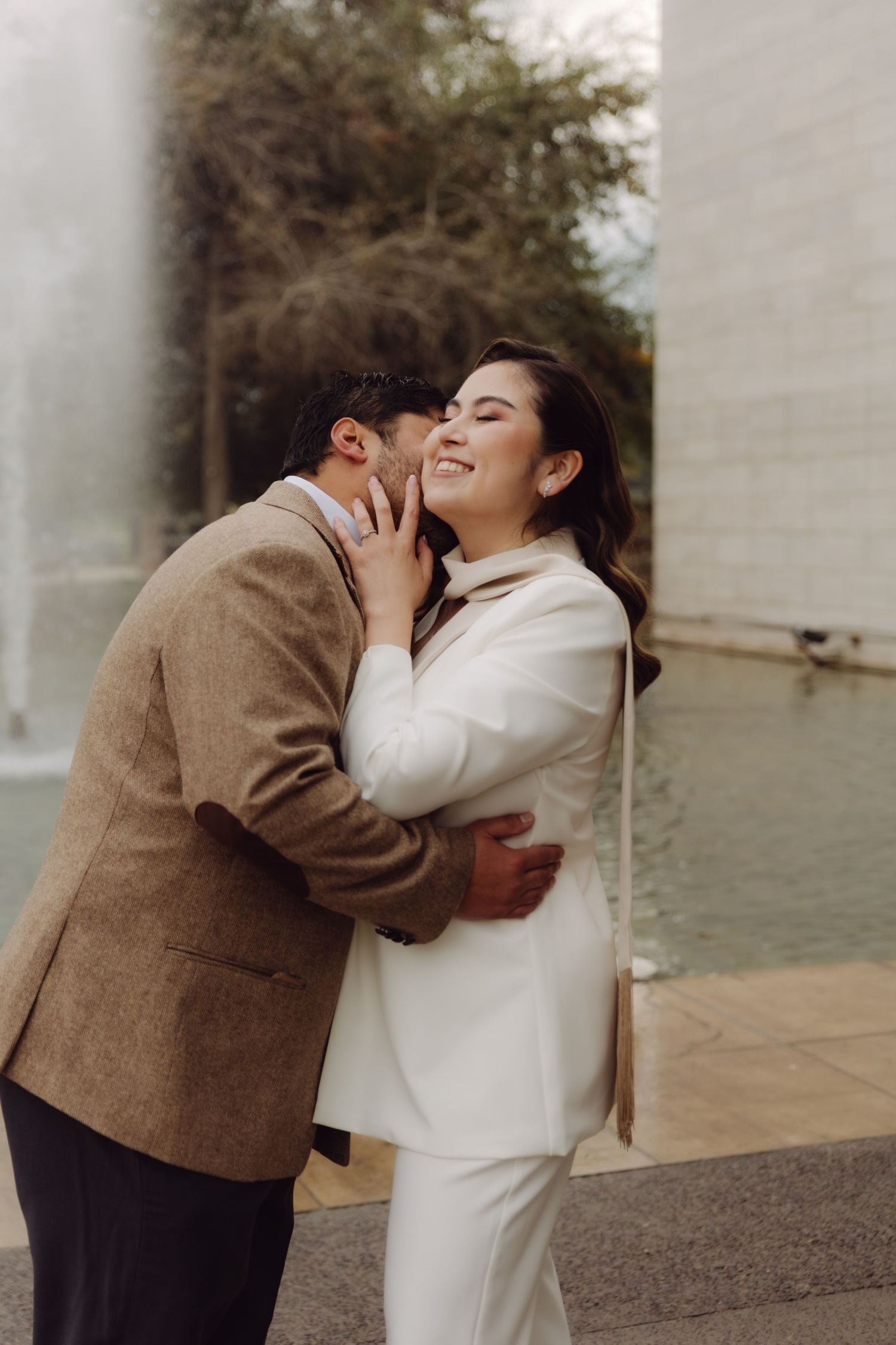 Couple sharing a romantic moment near a fountain, bride in white suit.