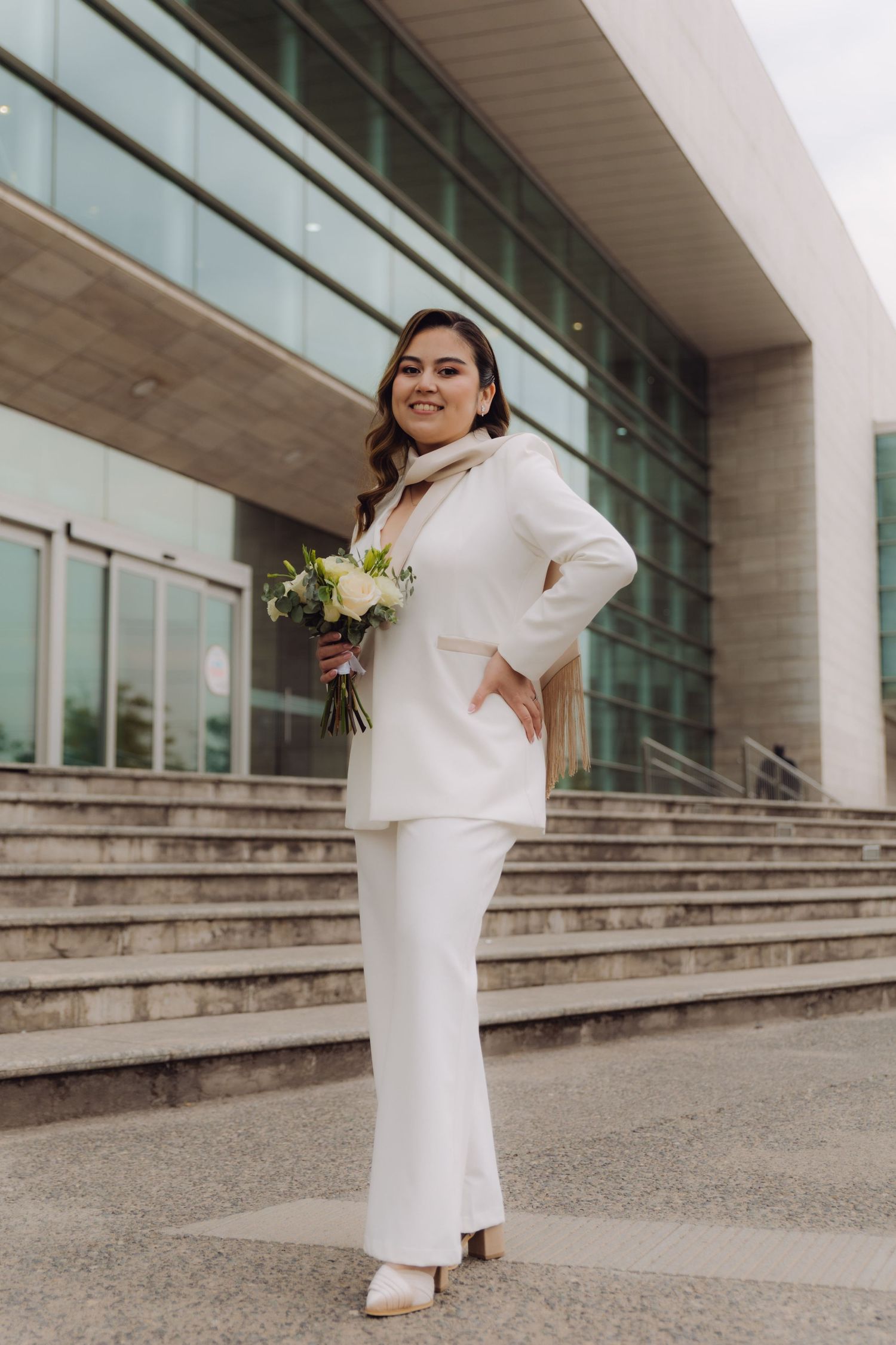 Bride in white pantsuit holding bouquet on steps of modern building.
