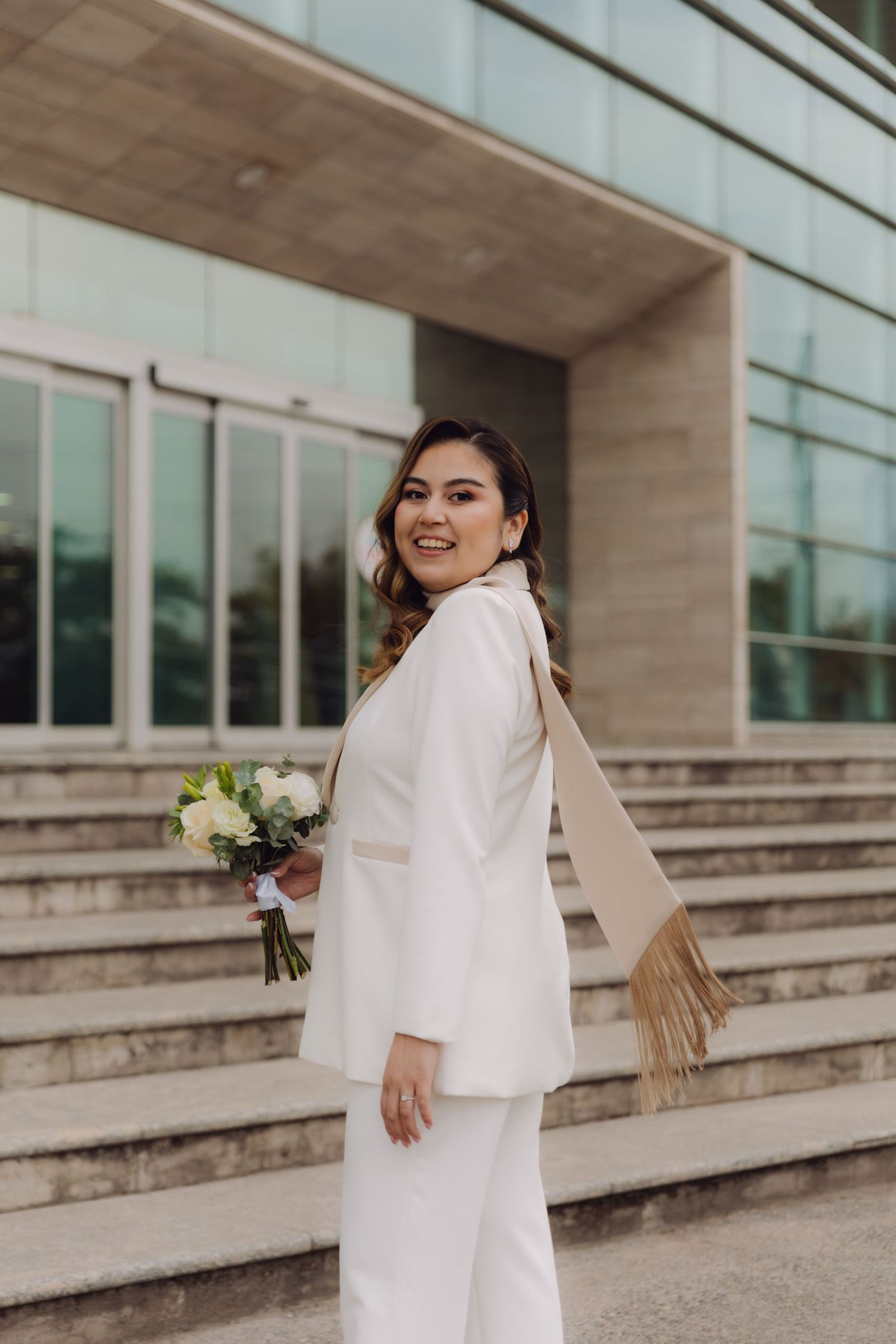 Smiling bride in white suit with fringe scarf holding bouquet on steps.