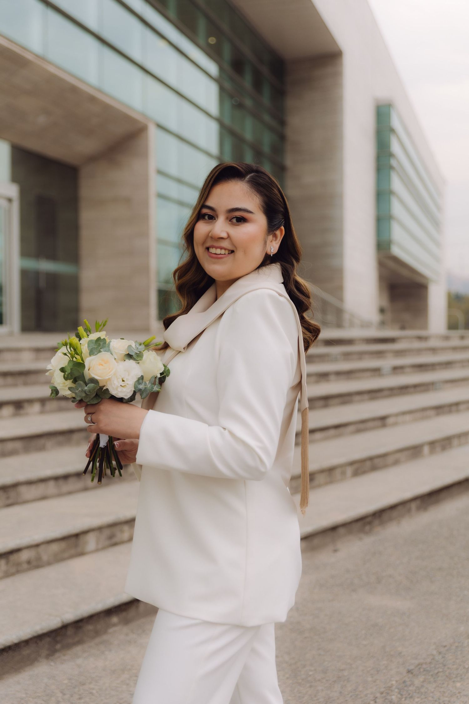 Elegant bride in white pantsuit holding white bouquet on stone steps.