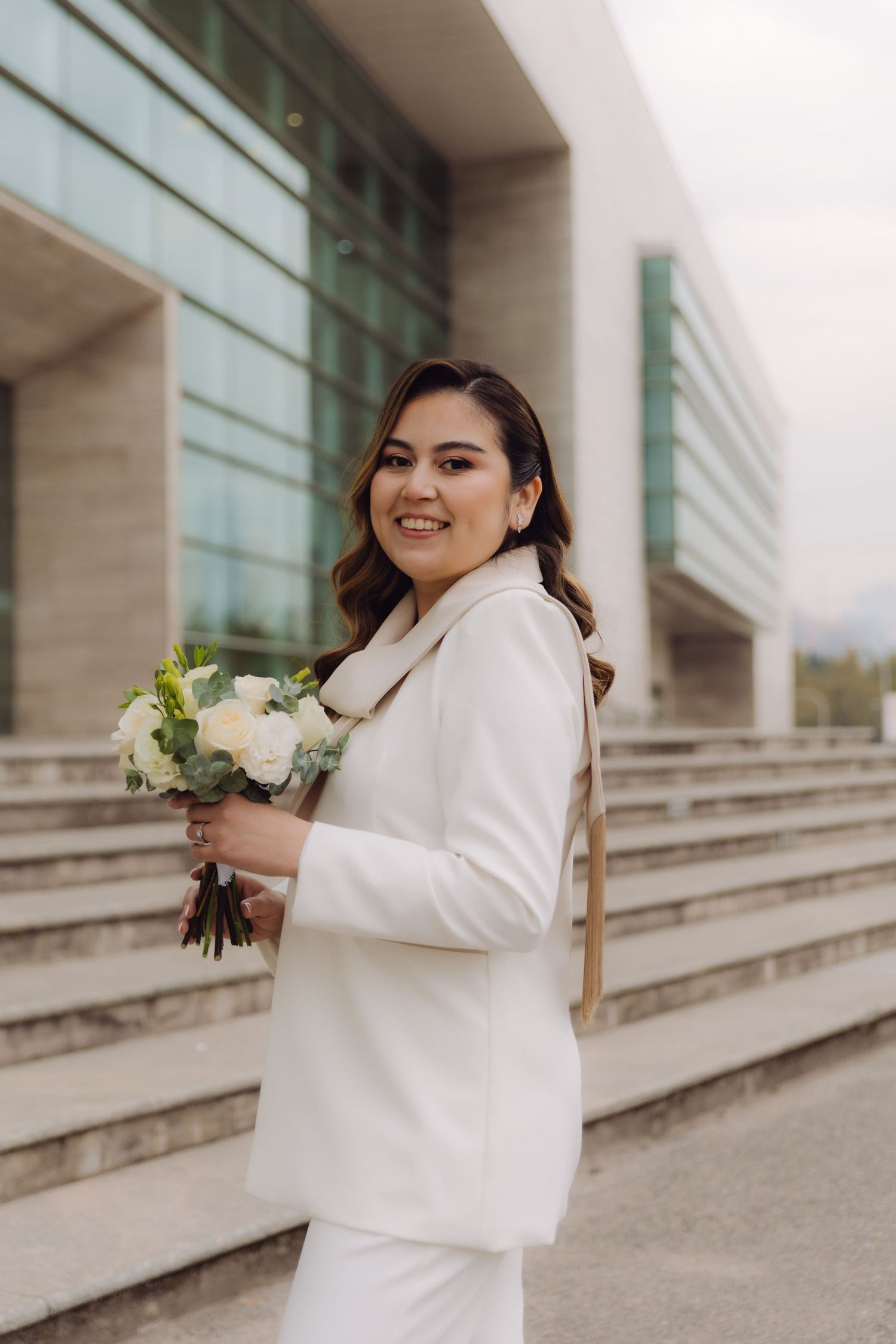 Radiant bride in white suit posing with bouquet outside modern building.