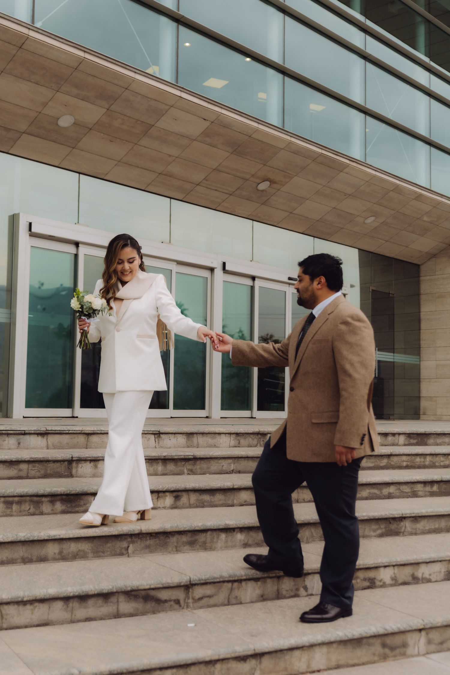 Bride in white suit holds groom's hand descending modern building steps, holding white bouquet.
