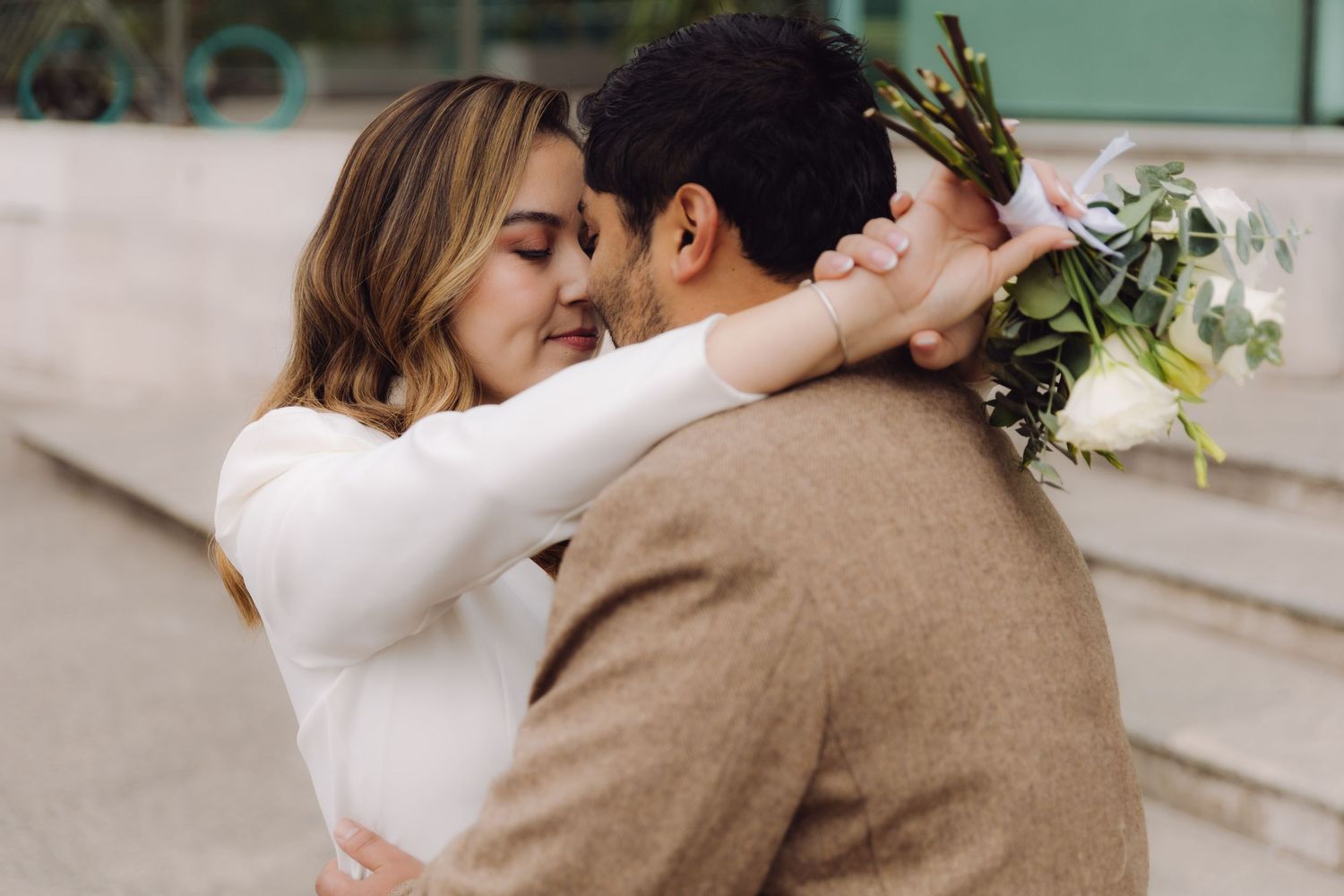 Couple sharing intimate moment, bride in white dress embracing groom in tan blazer with bouquet.