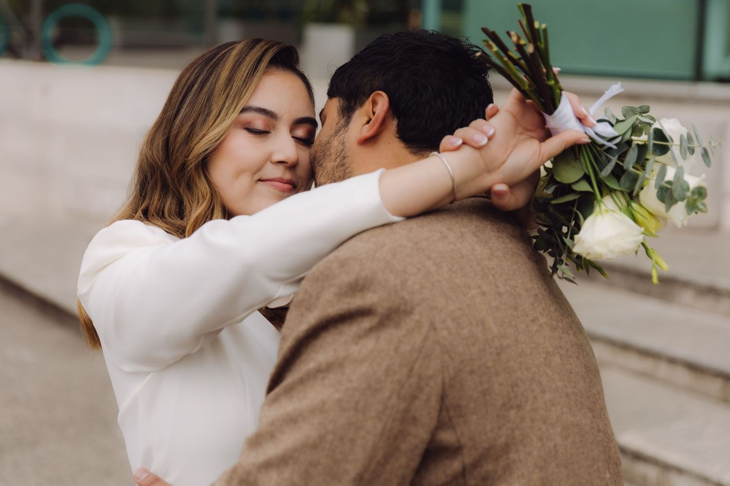 Smiling bride in white outfit embraces groom in brown jacket while holding white floral bouquet.