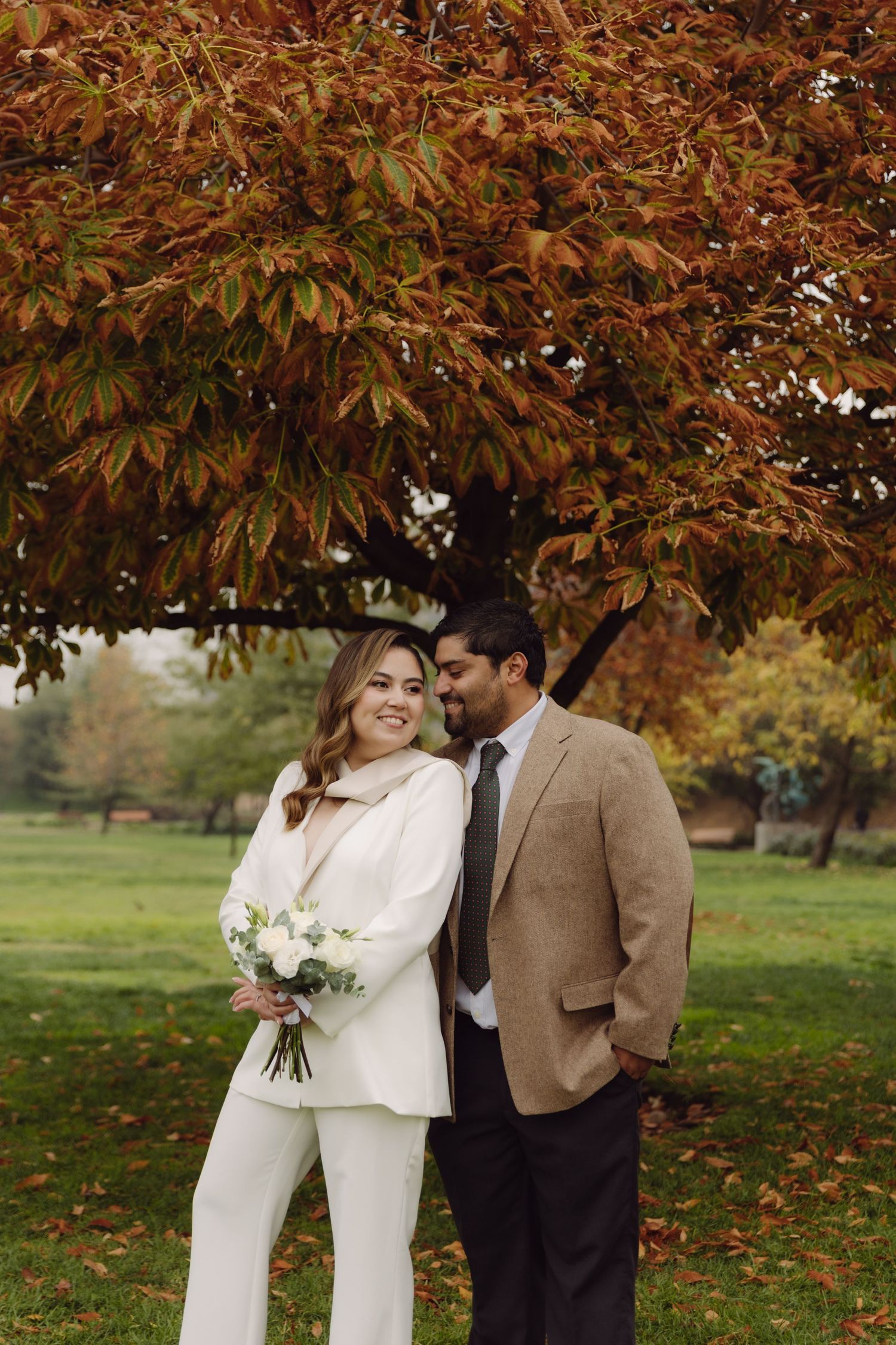 Bride in white pantsuit holding bouquet smiles beside groom in tan blazer under autumn tree.