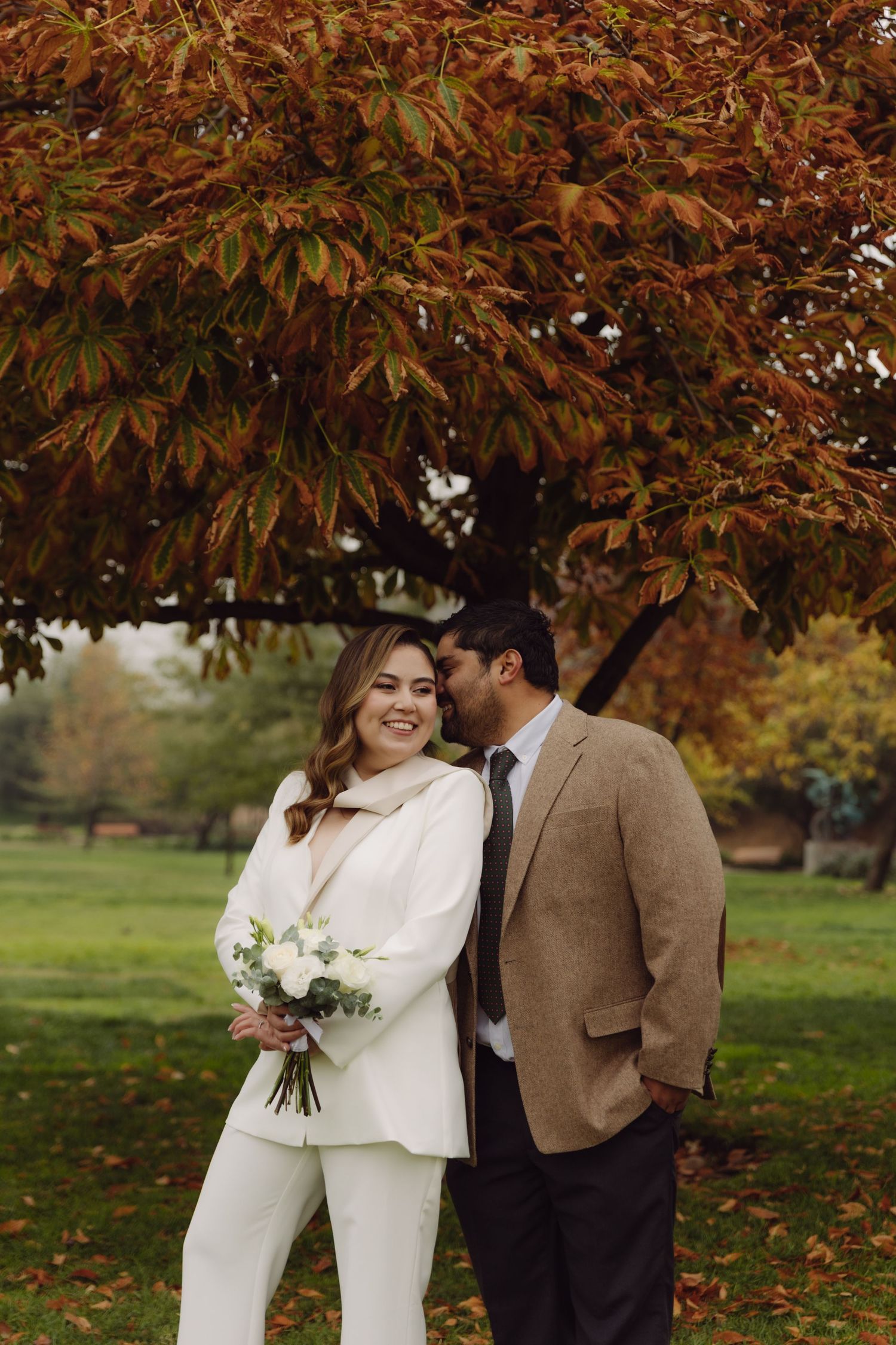 Groom in tan blazer whispers to laughing bride in white suit under vibrant fall foliage.