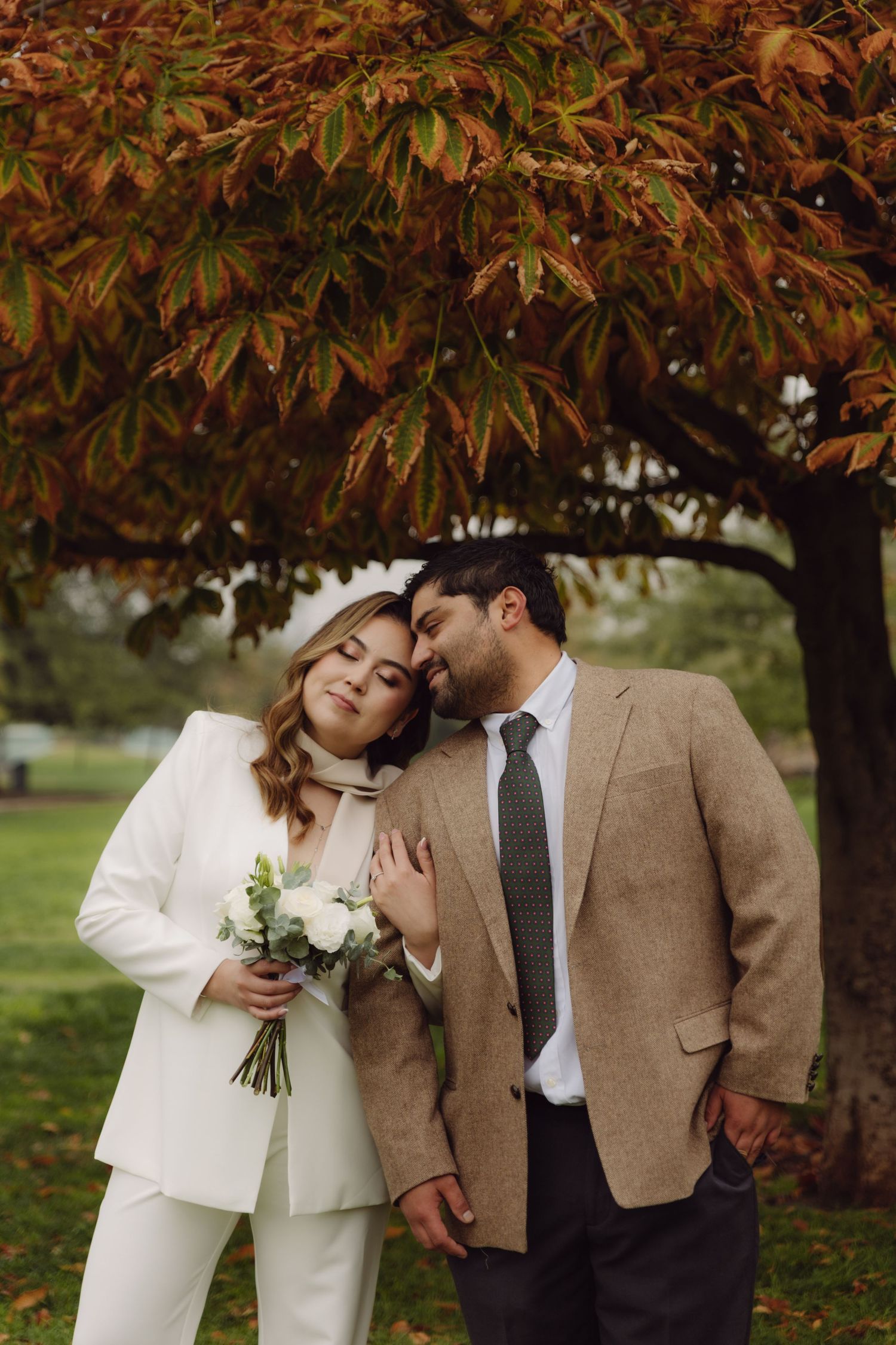 Wedding couple leans together tenderly under red autumn tree, bride holding white rose bouquet.
